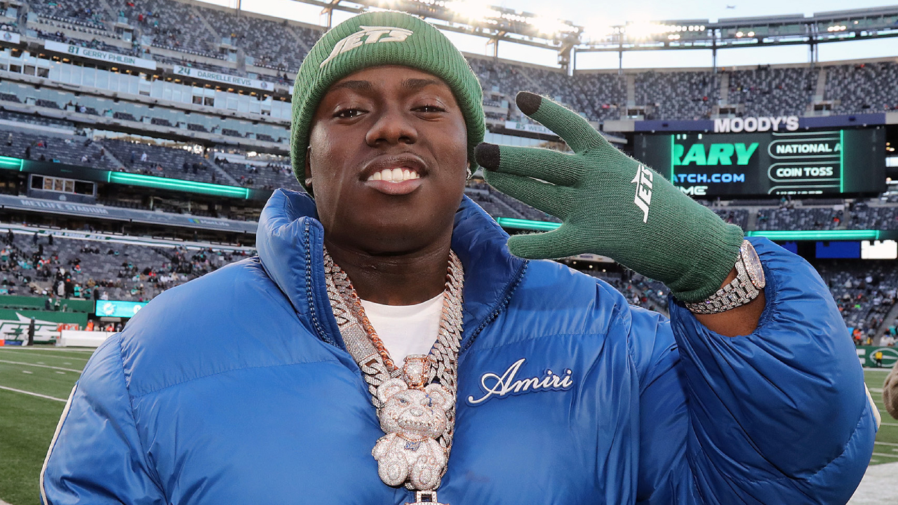 Sheff G in a blue jacket and green beanie, wearing large chains, smiles and gestures with a gloved hand at a stadium.