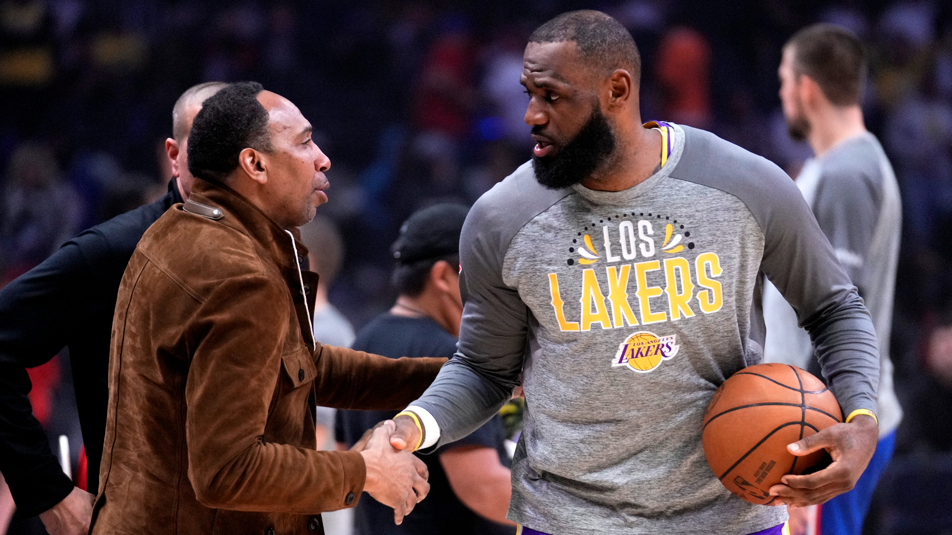 LeBron James in a Lakers shirt shakes hands with Stephen A. Smith, who is wearing a brown jacket, on a basketball court.
