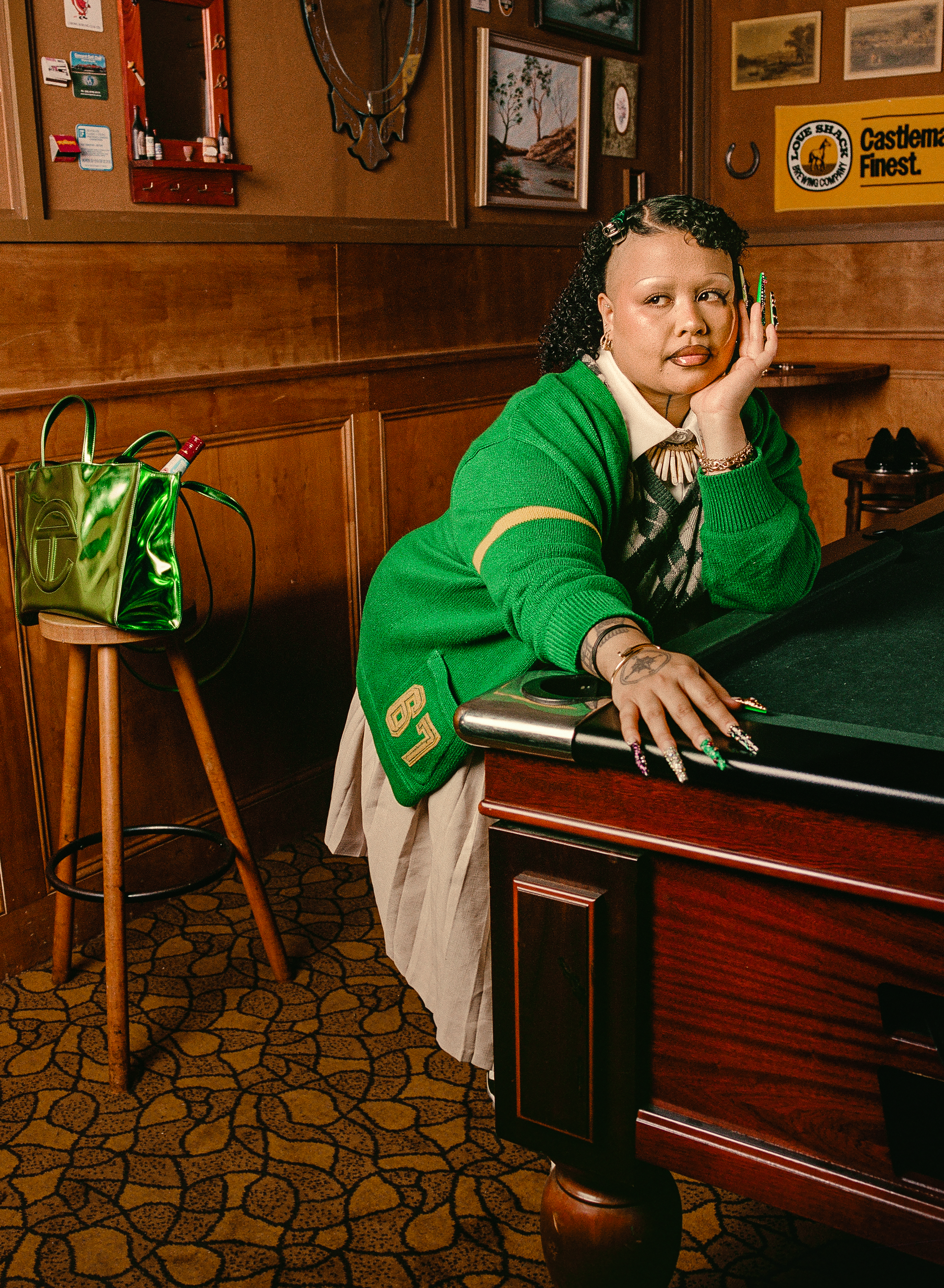 A person in a green sweater leans on a pool table in a vintage-style room. A shiny green bag sits on a stool nearby.