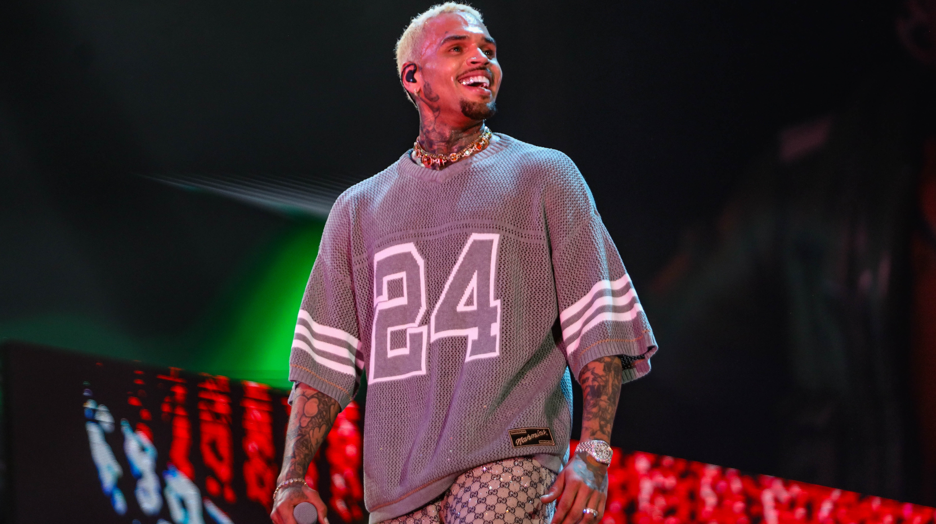 Chris Brown with blond hair and tattoos performs on stage, wearing a gray jersey with the number 24 and patterned pants.