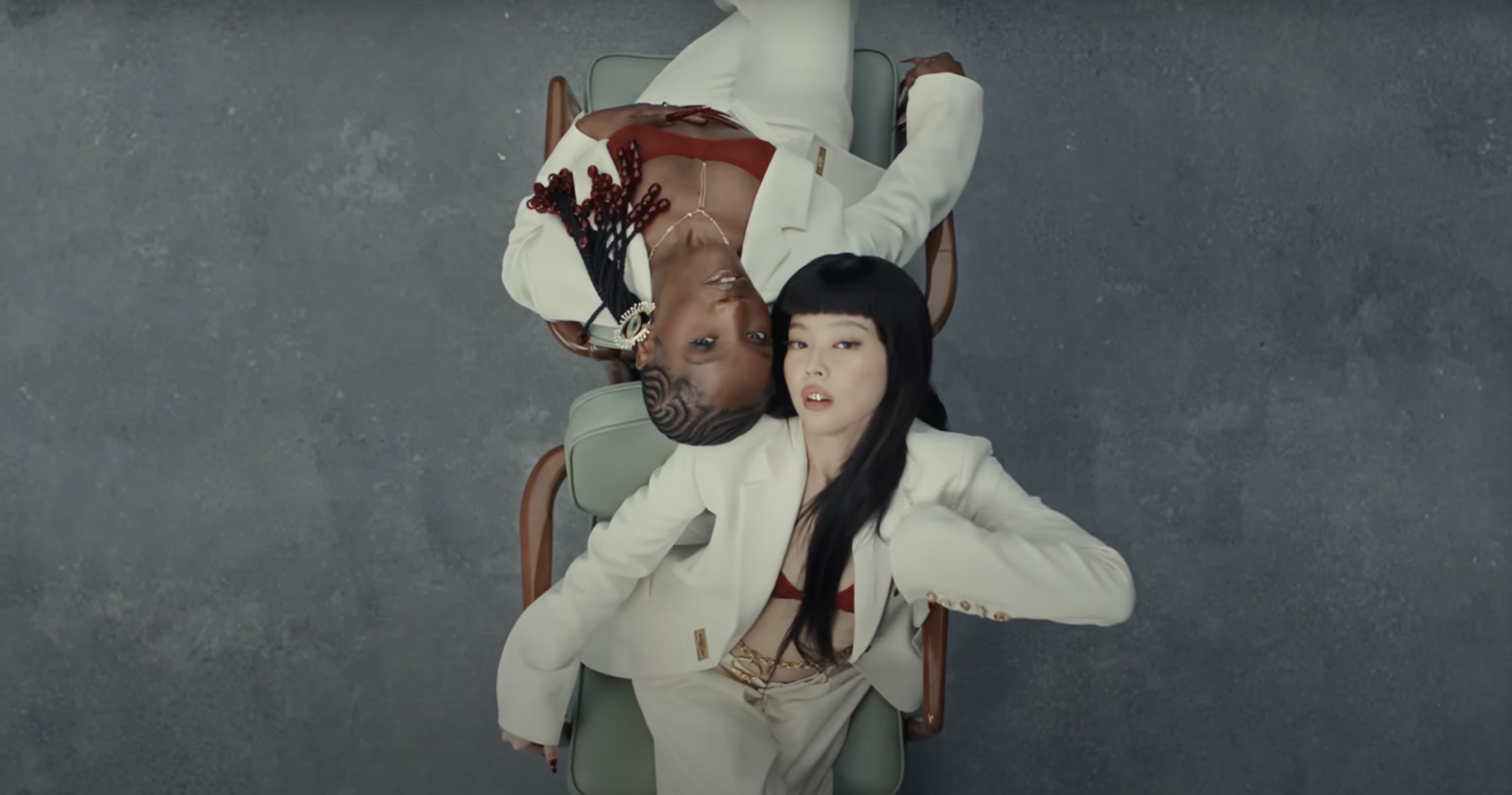 Jennie and Doechii in stylish white outfits lie on a chair, one with braided hair and the other with long black hair, against a gray background.