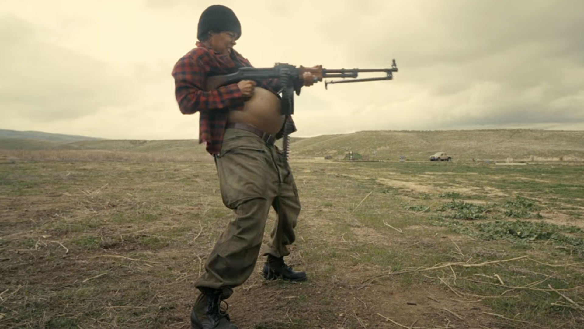 Teyana Taylor in a field firing a rifle, wearing a plaid shirt, beanie, and boots, with an open landscape in the background.