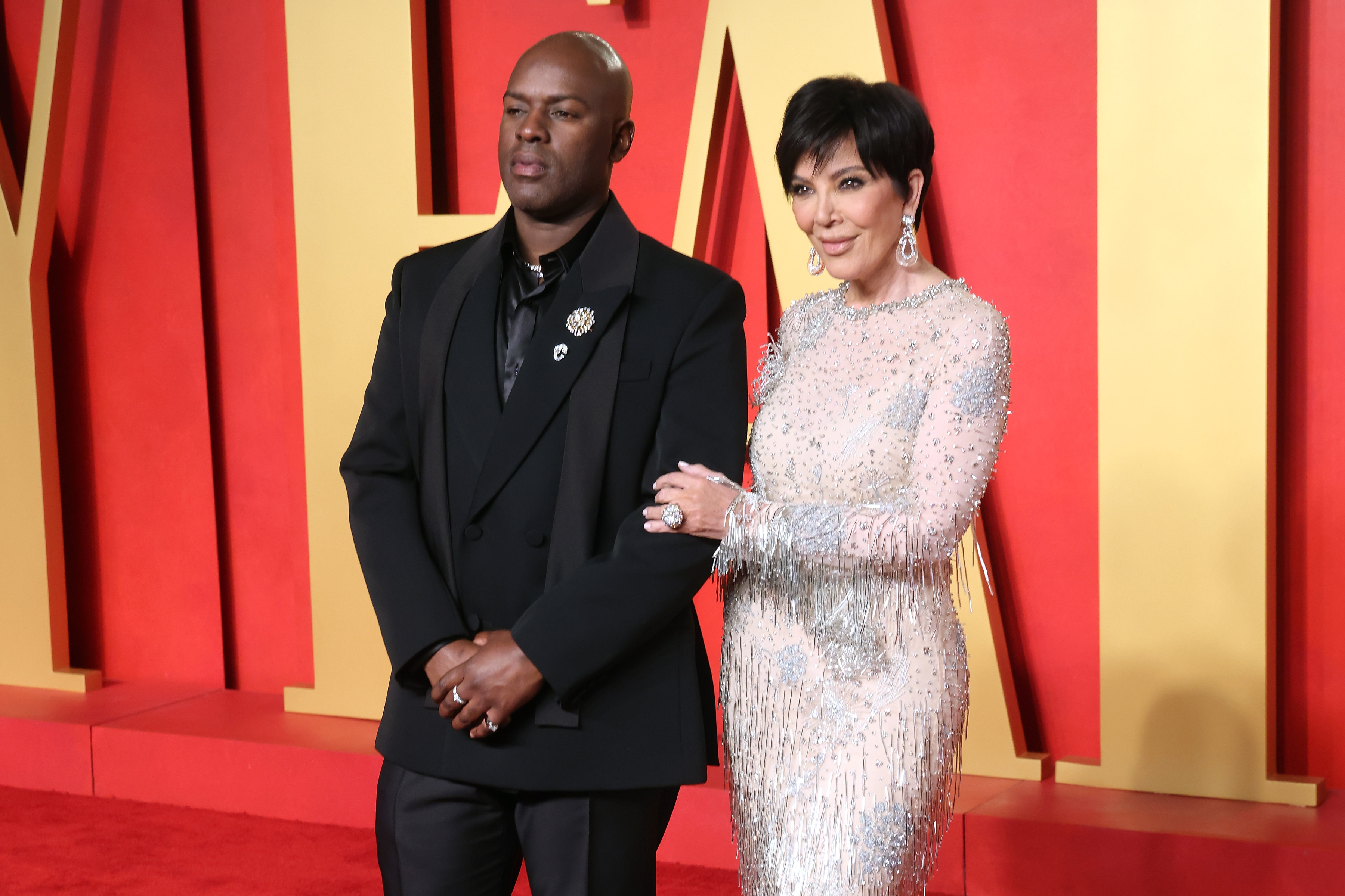 Kris Jenner in a shimmering dress poses with Corey Gamble in a black suit on a red carpet with a red and yellow backdrop.