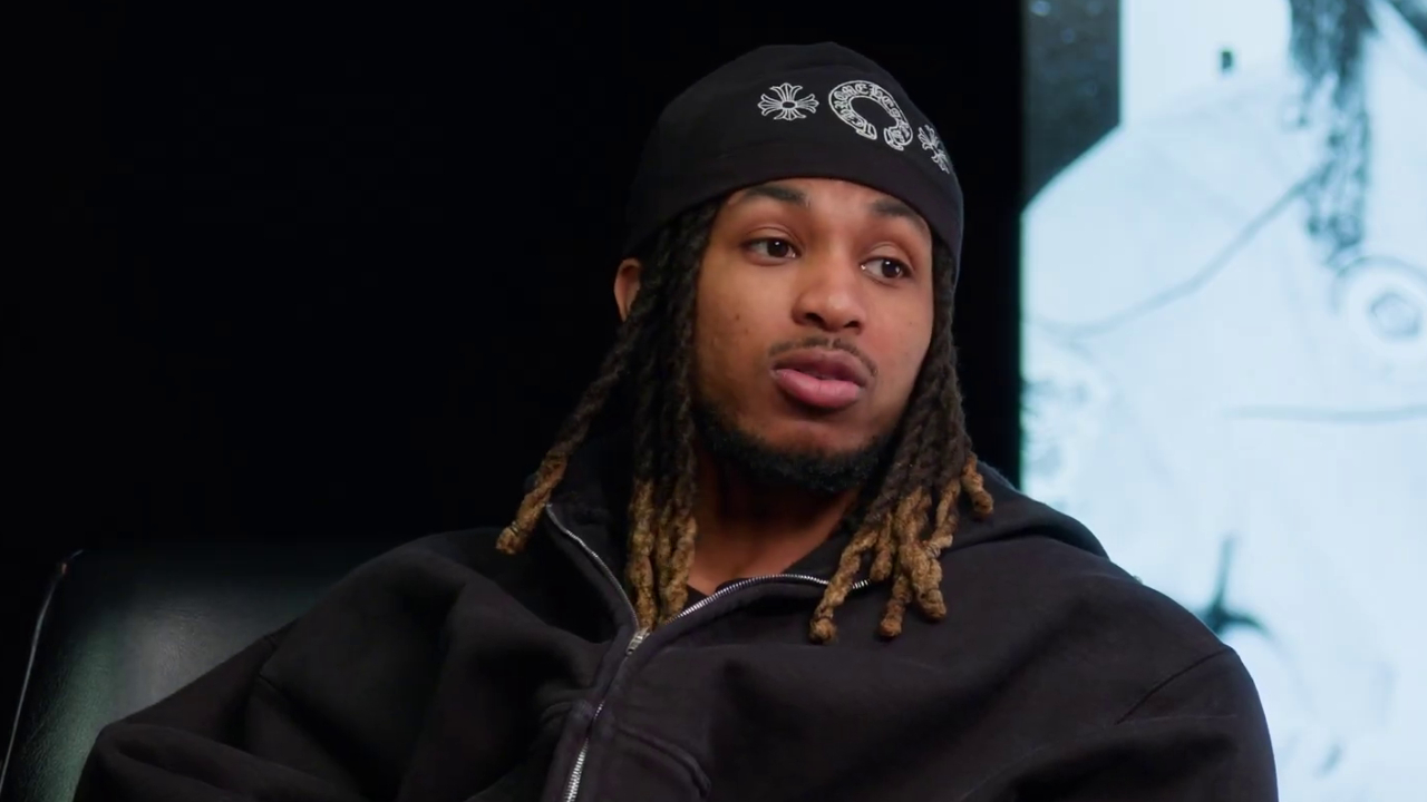 DDG with long braids and a black bandana is sitting in a dark setting, wearing a black hoodie.