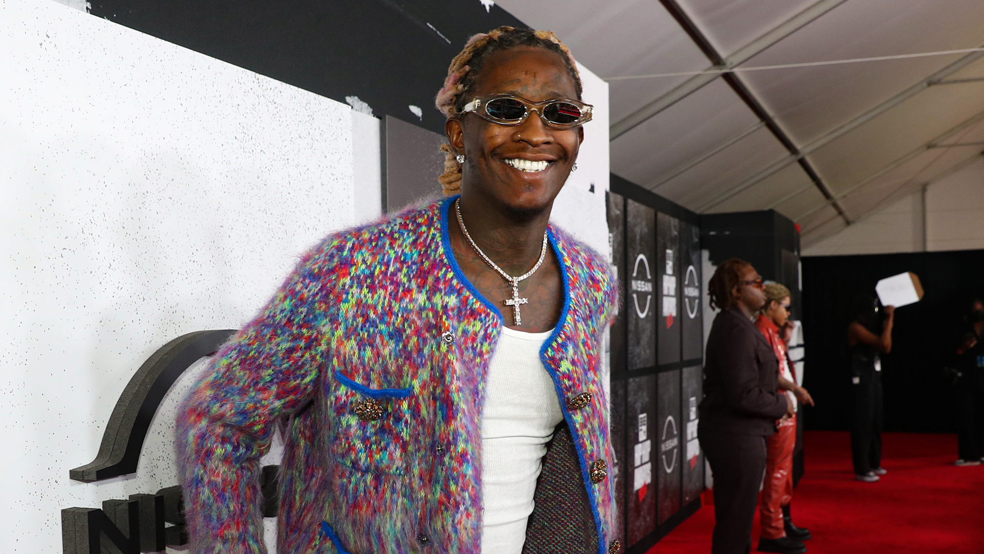 Young Thug wearing a colorful jacket and sunglasses smiles on the red carpet at an event, with other people in the background.