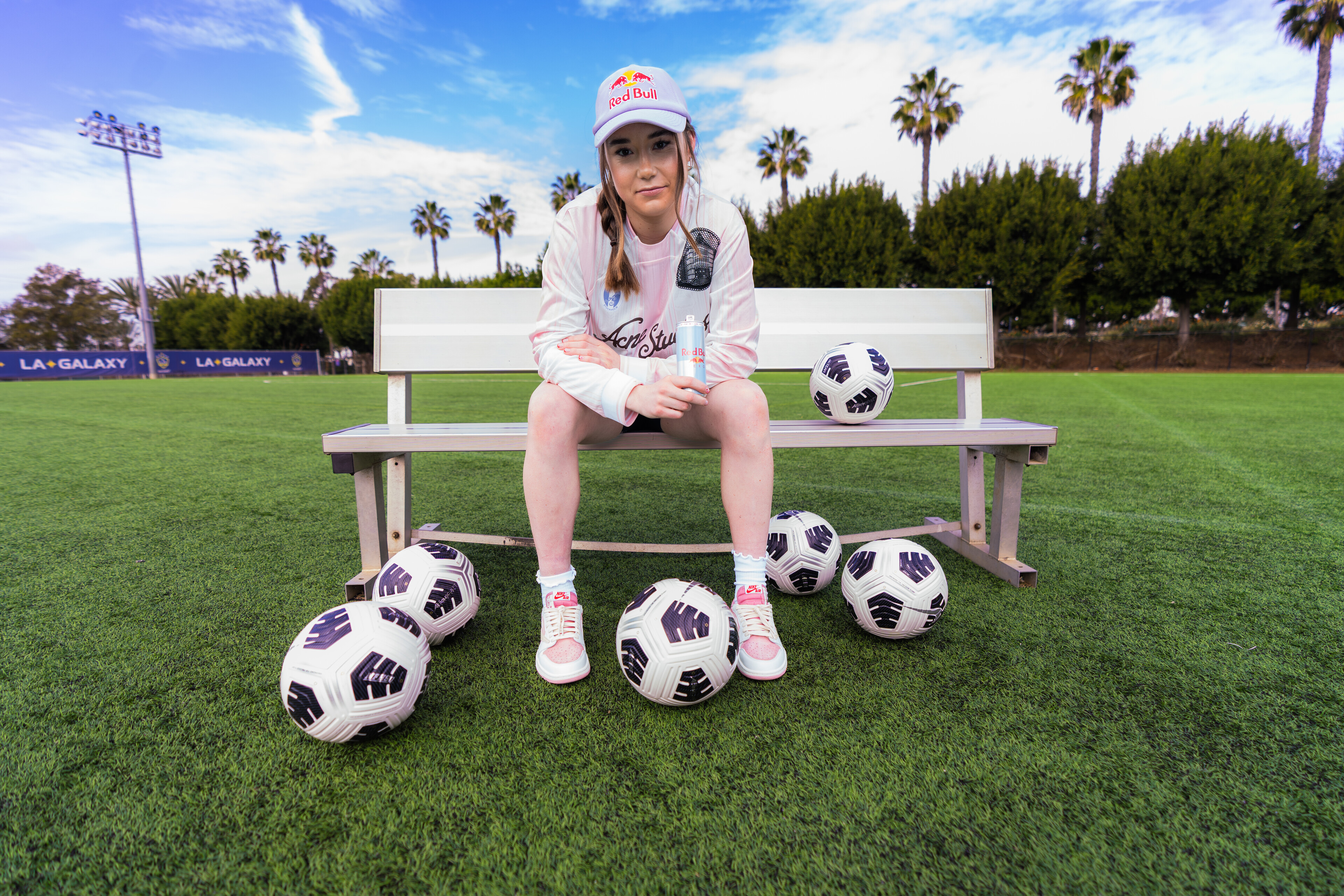 Olivia Moultrie wearing a Red Bull cap sits on a bench surrounded by soccer balls on a field, with palm trees and a blue sky in the background.