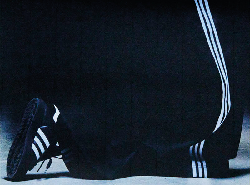 A person wearing black Adidas sneakers and track pants with white stripes, kneeling on the ground.