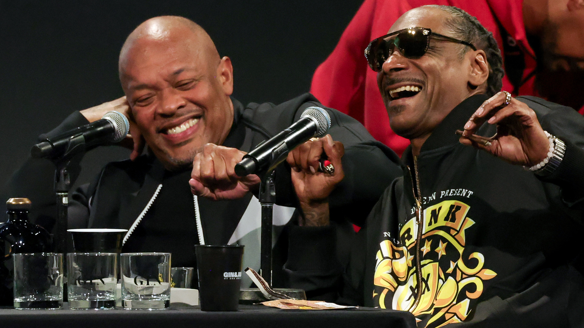 Dr. Dre and Snoop Dogg laughing at a panel, both wearing dark clothing. Dr. Dre is bald, and Snoop Dogg has braided hair and sunglasses.