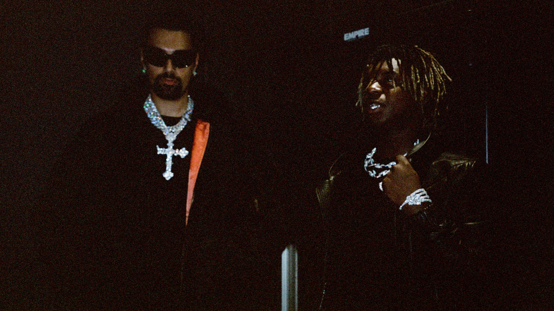 Two individuals in dark clothing and jewelry, one wearing sunglasses. The setting is dimly lit.