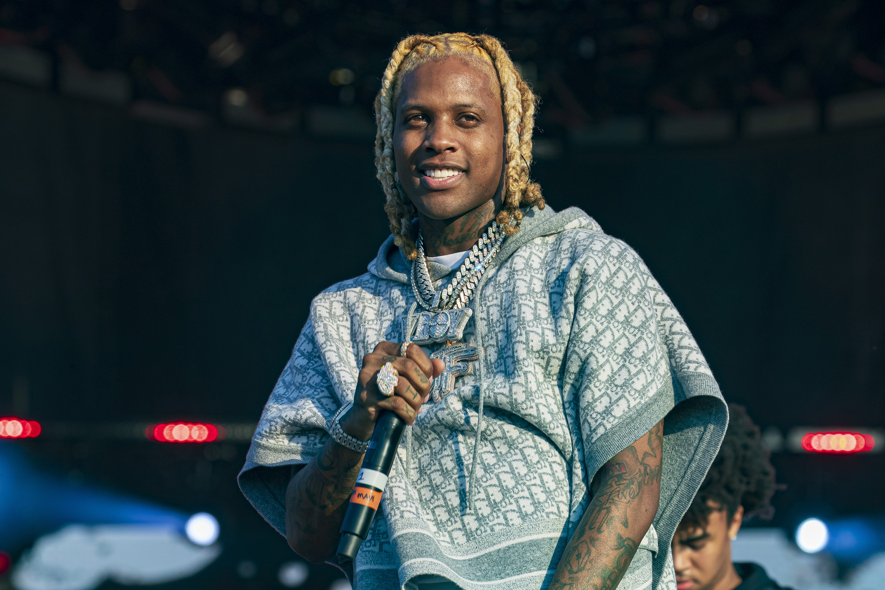 Rapper Lil Durk performing on stage, wearing a patterned hoodie and jewelry, with a microphone in hand.