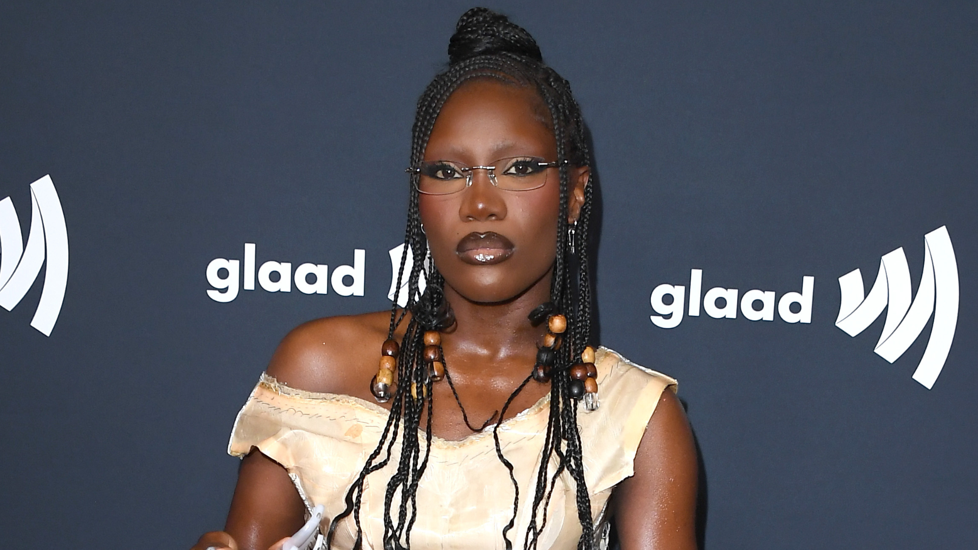 A person with braided hair and glasses poses in front of a GLAAD backdrop, wearing a beige outfit with bead details.