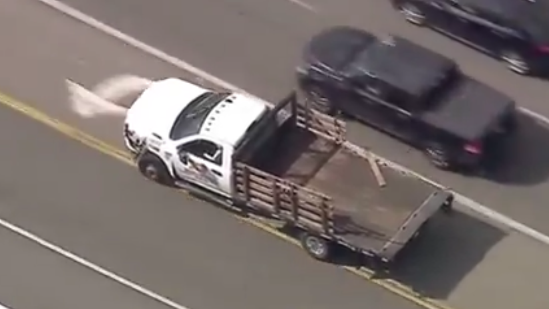 Aerial view of a white flatbed truck driving on a road, with a black vehicle nearby. The truck has wooden side rails.