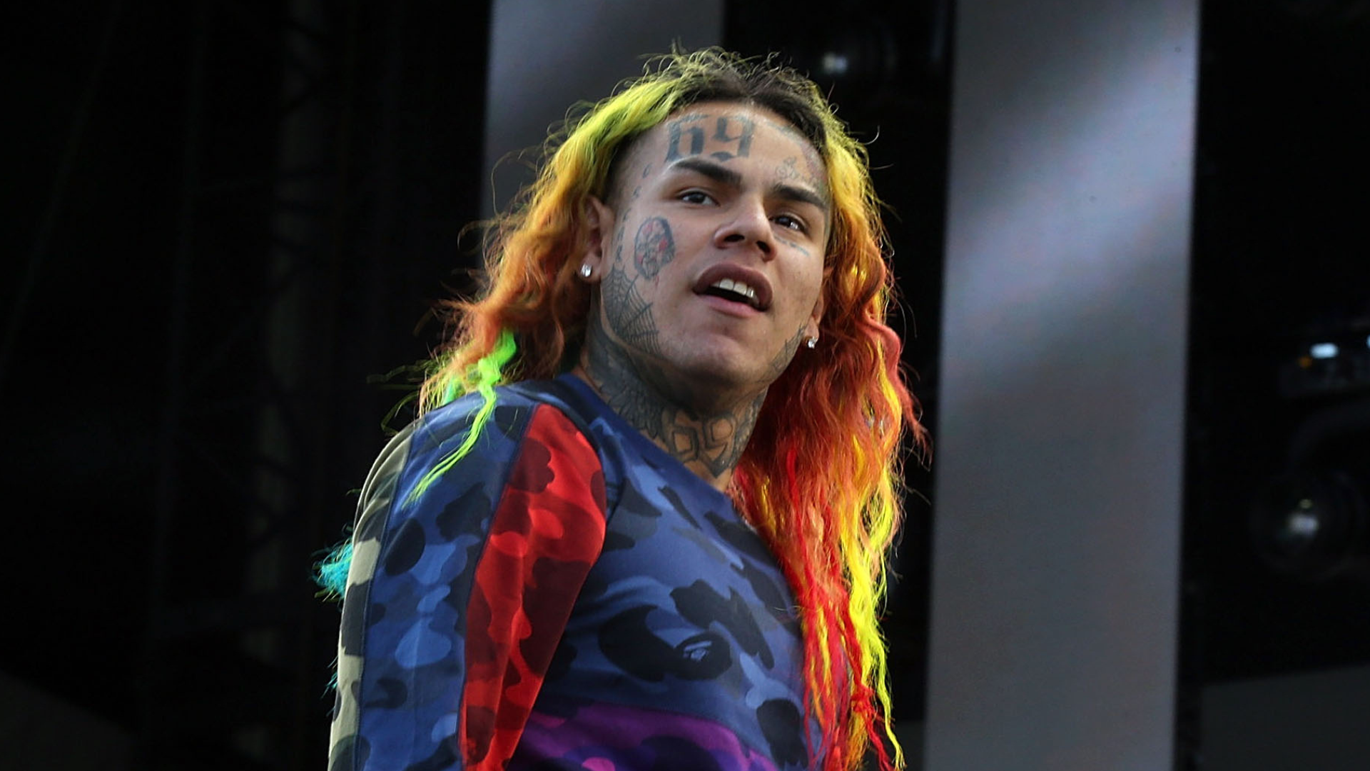 Rapper Tekashi 6ix9ine with rainbow hair and tattoos, wearing a colorful shirt, performing on stage.