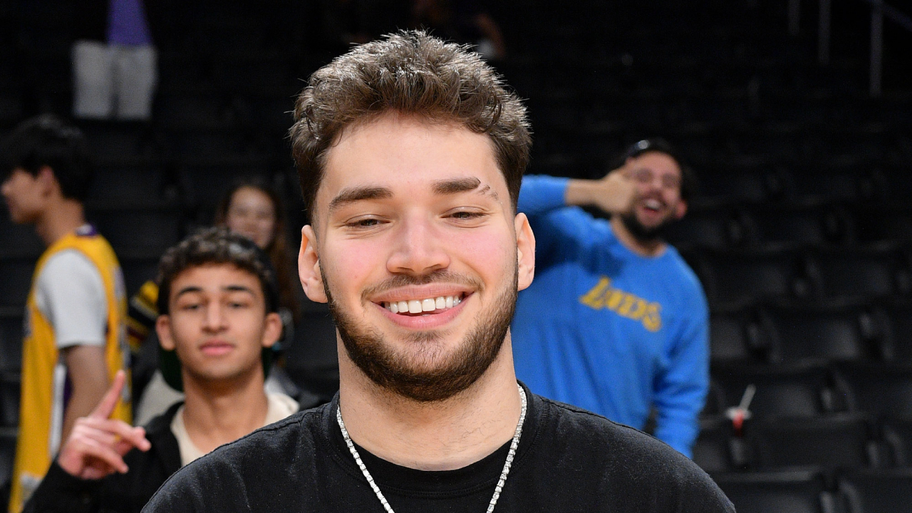 Adin Ross smiles with short hair and a beard in the foreground, with people in the background, one wearing a Lakers shirt.
