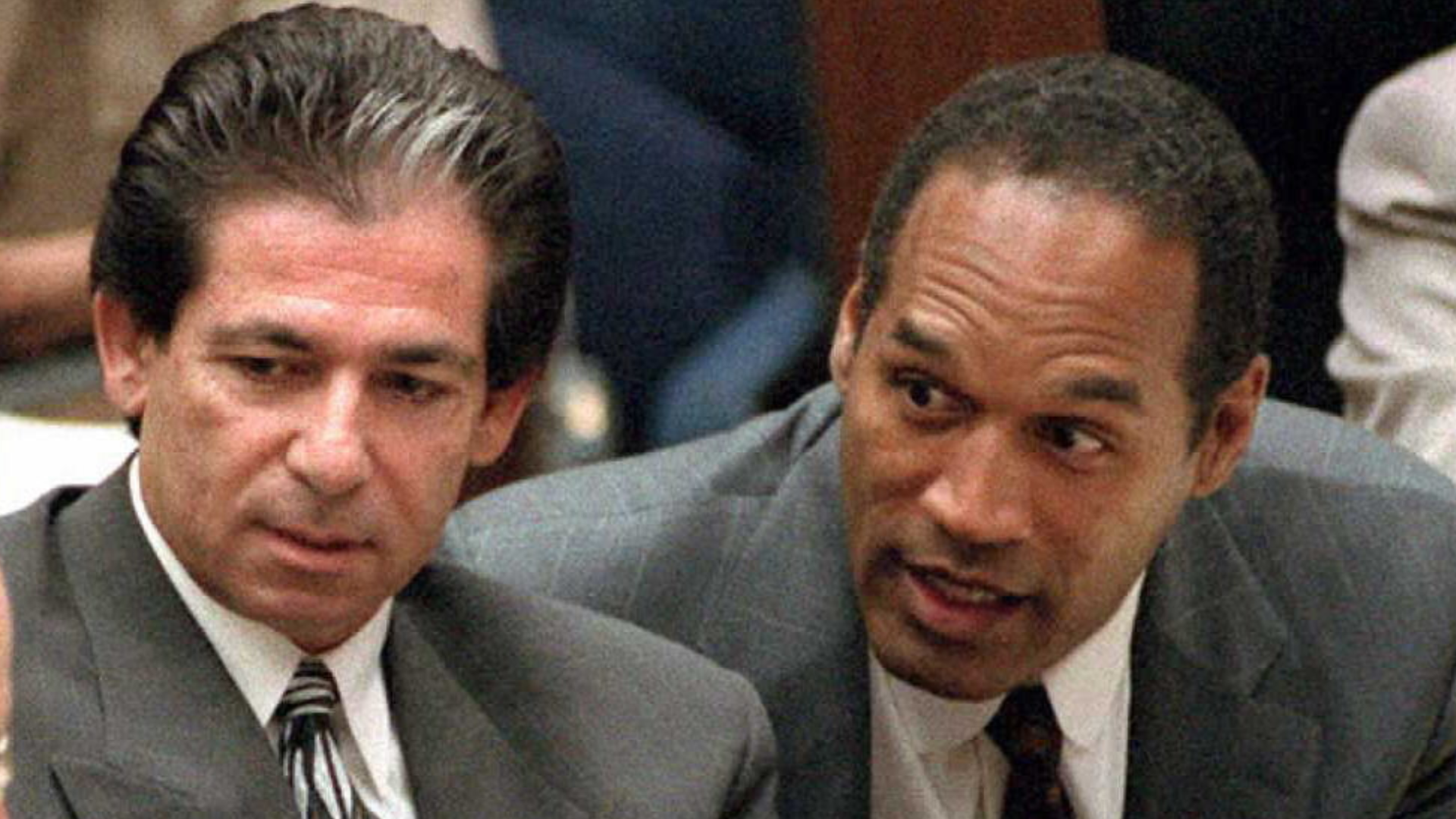 Robert Kardashian and O.J. Simpson in a courtroom, both wearing suits and engaged in conversation.