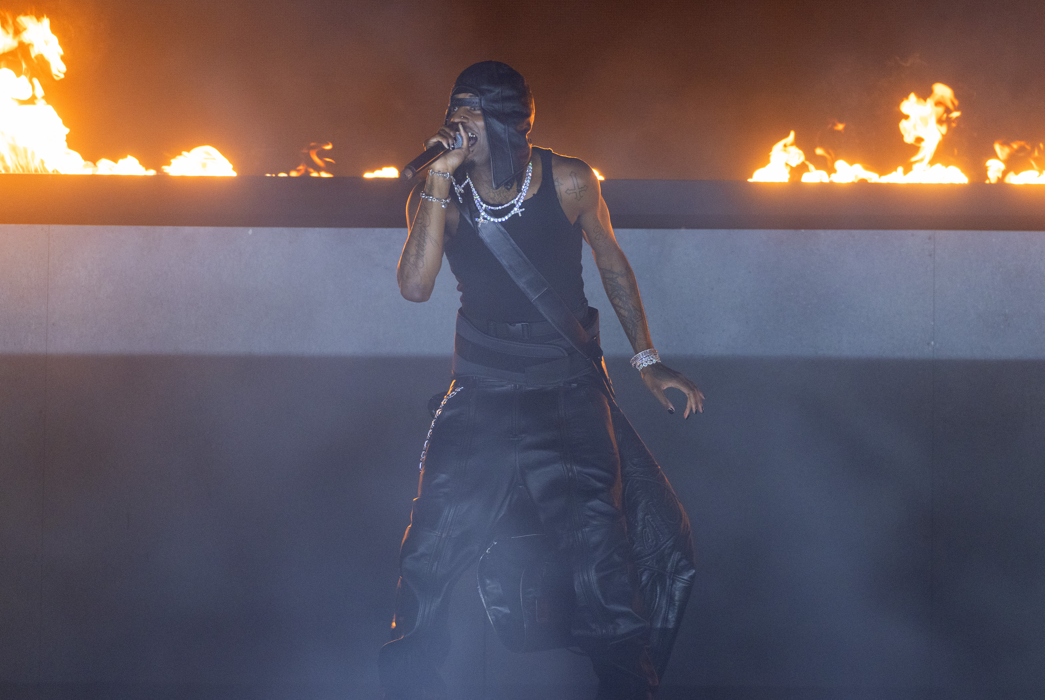 Playboi Carti in dark clothing and a cap sings on stage with flames in the background, creating a dramatic and intense atmosphere.
