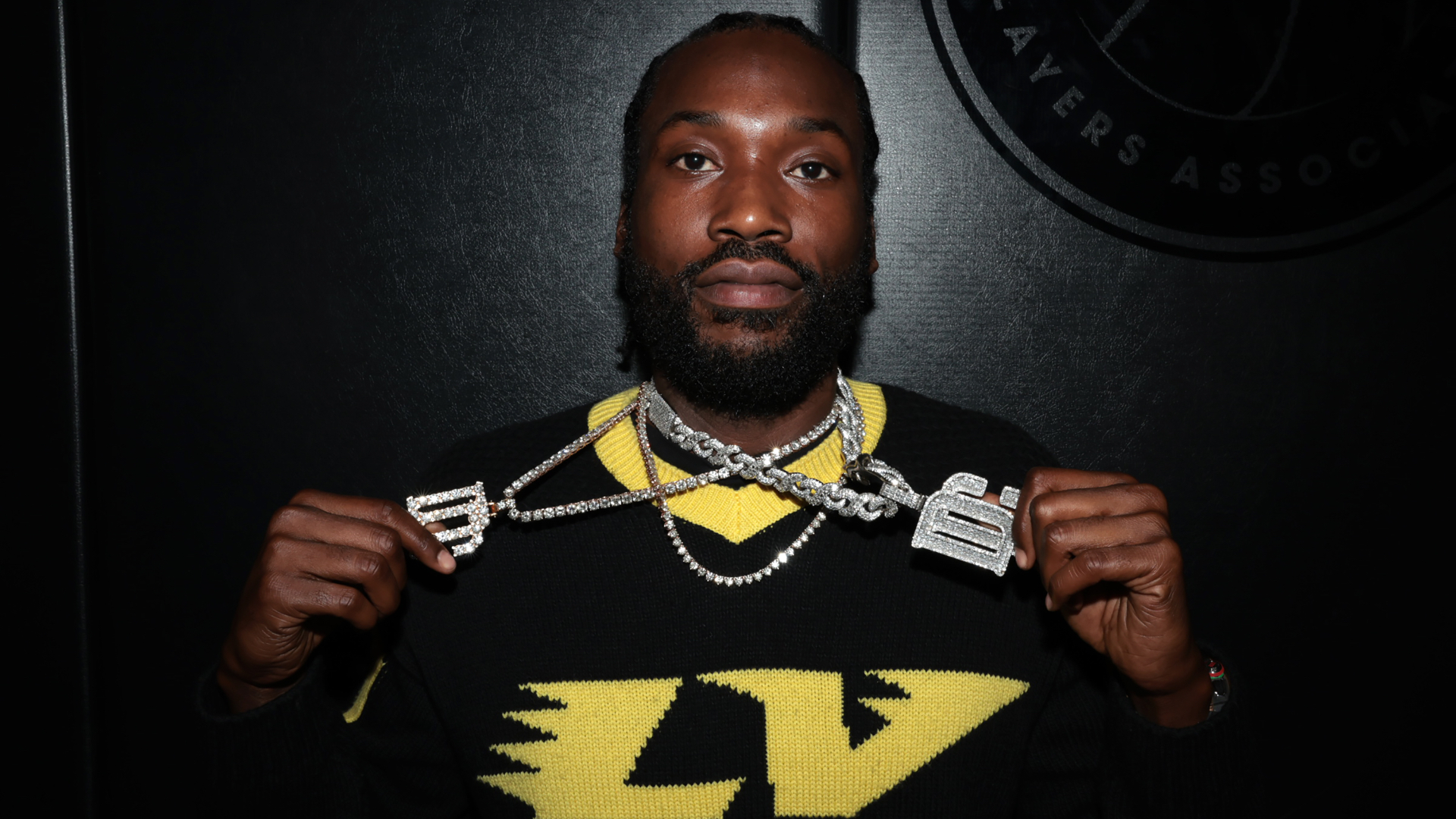 Meek Mill holding two large chains with pendant letters, wearing a black and yellow sweater, against a dark background.