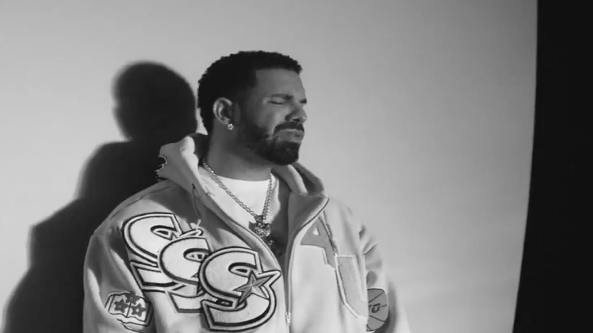A black and white photo of Drake wearing a jacket with large letter designs, standing against a plain background.