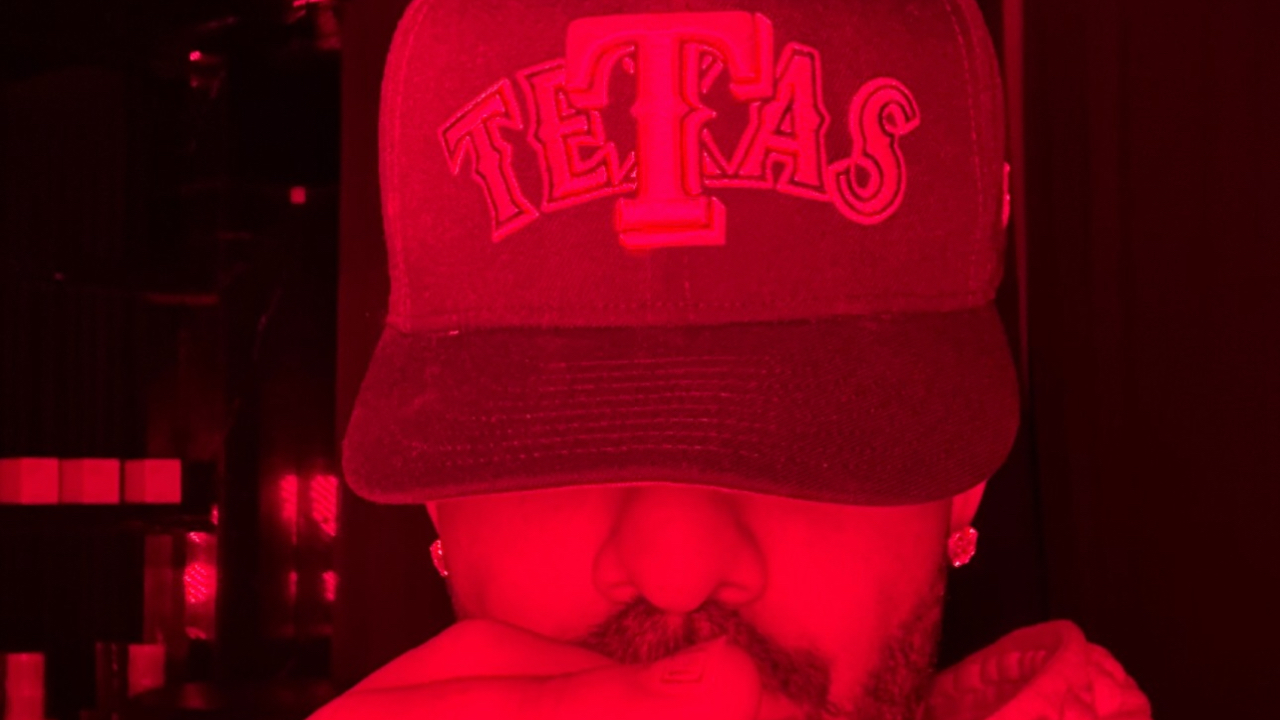 Drake wearing a cap with "TETAS" text, partially covering his face, in red lighting.