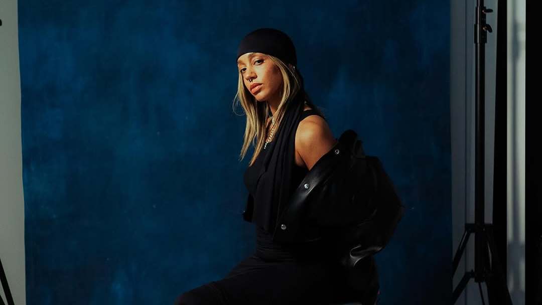 Martine Ali in a black outfit and headscarf poses against a blue backdrop, with studio lighting equipment visible.