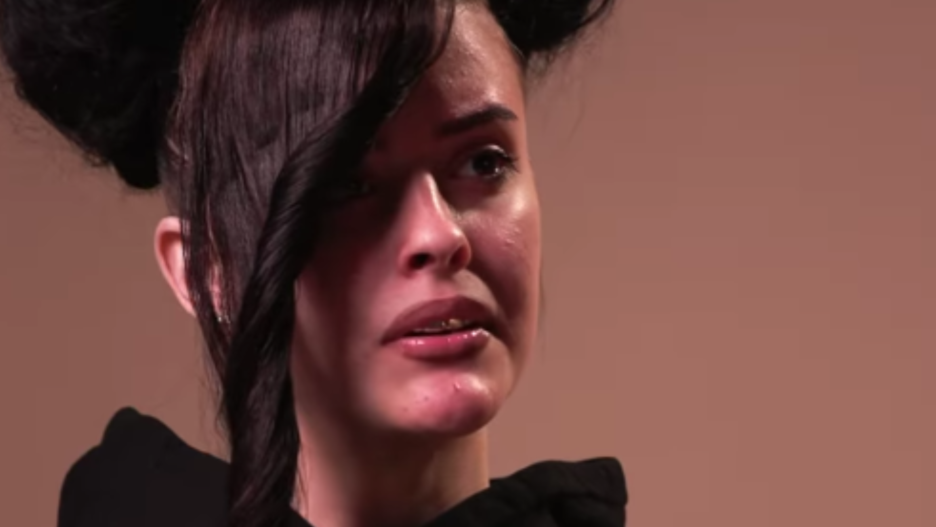 A person with a unique hairstyle and an emotional expression, wearing a dark outfit, against a neutral background.