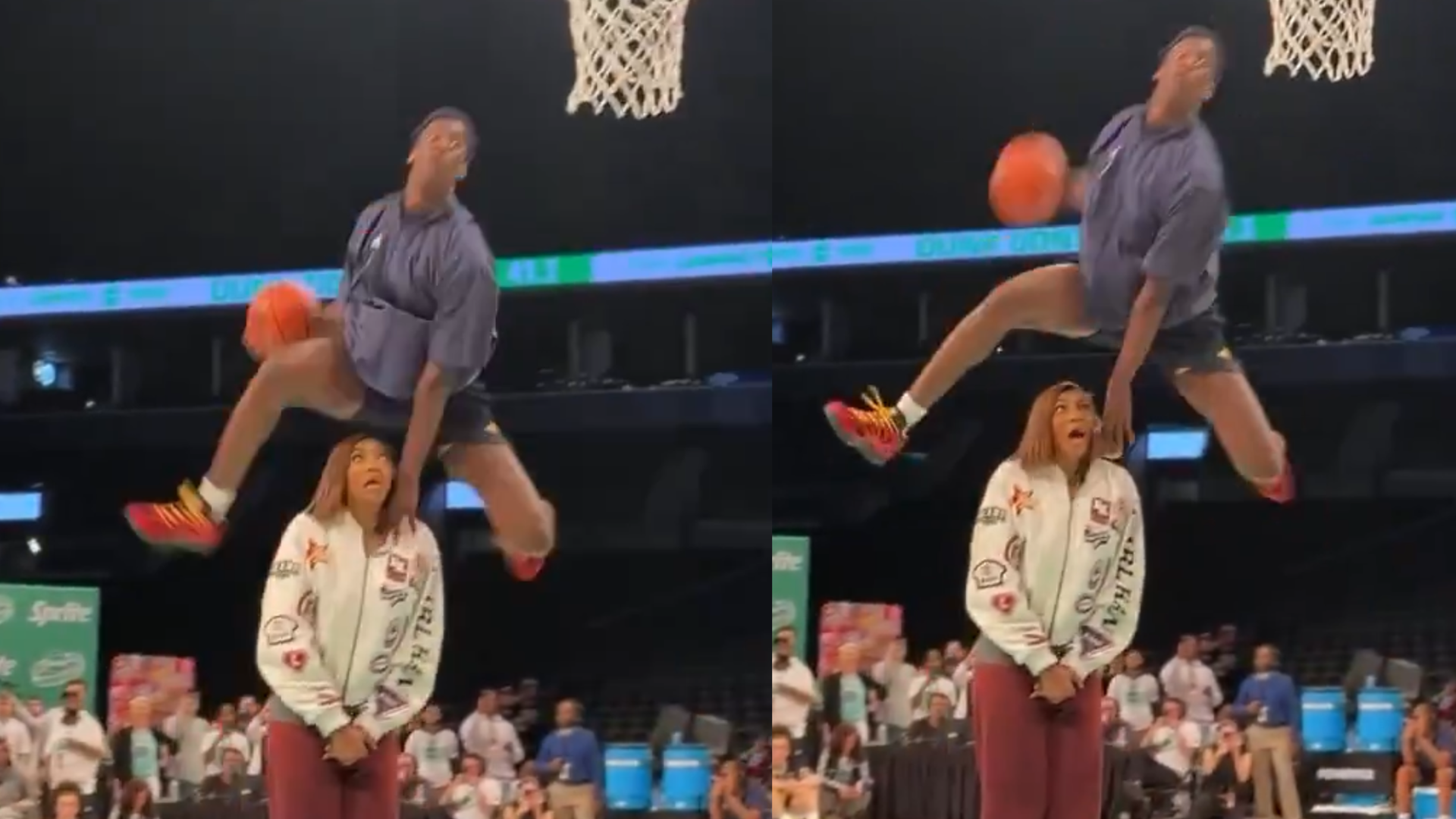 A basketball player performs a slam dunk over a woman standing below the hoop in an indoor arena.