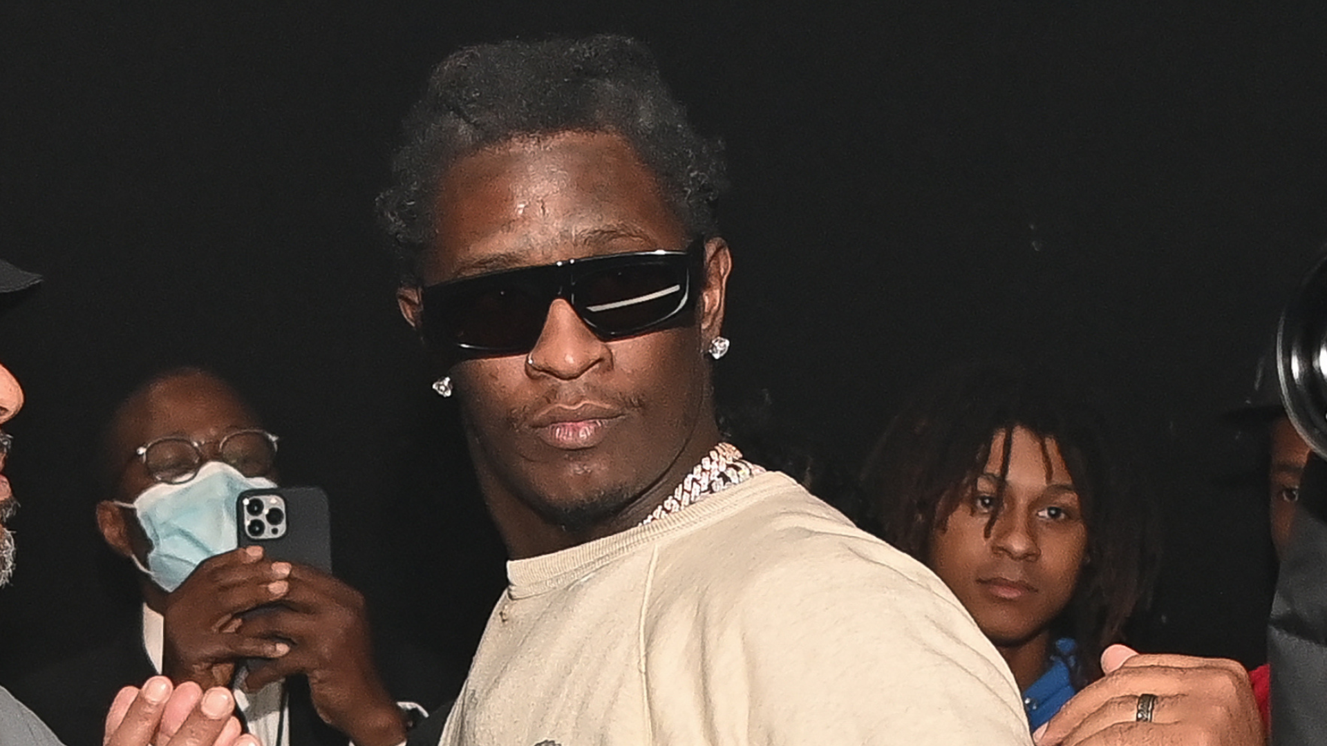 Young Thug wearing sunglasses and a necklace in a dimly lit setting, surrounded by people.