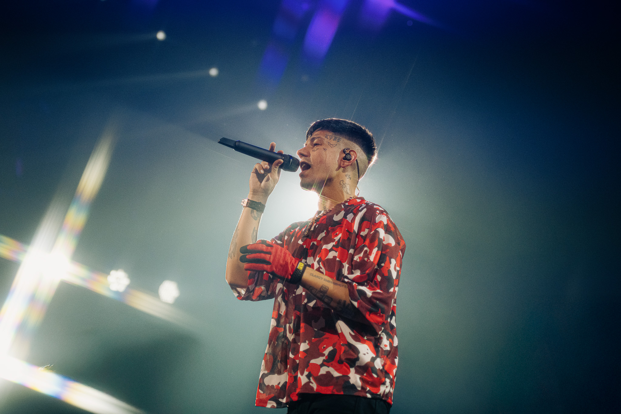 Duki performing on stage, wearing a red camouflage shirt and red gloves, holding a microphone with bright stage lights behind.