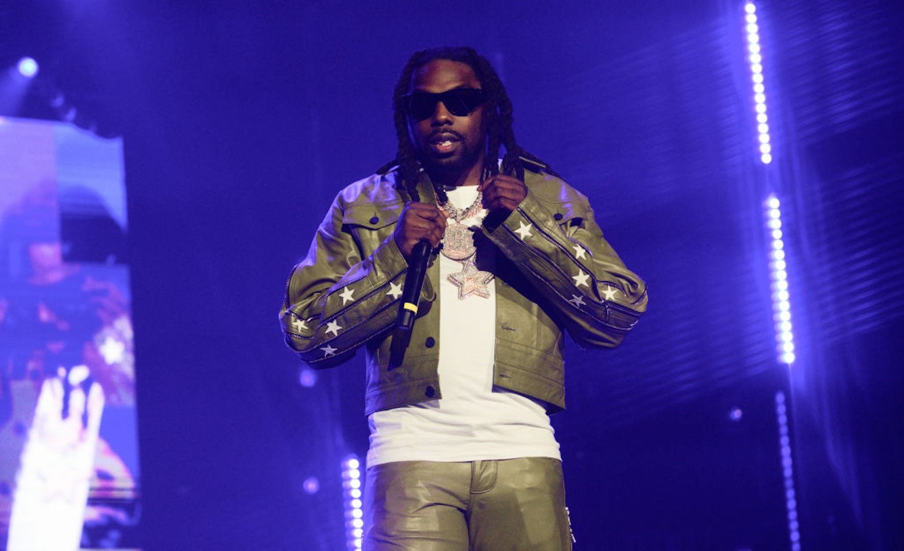 A man in a green leather outfit with star patterns performs on stage, wearing sunglasses and holding a microphone.