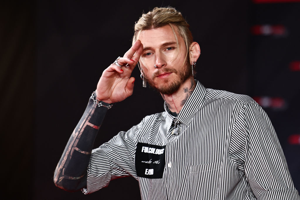 Machine Gun Kelly saluting on the red carpet, wearing a striped shirt and showcasing tattoos.