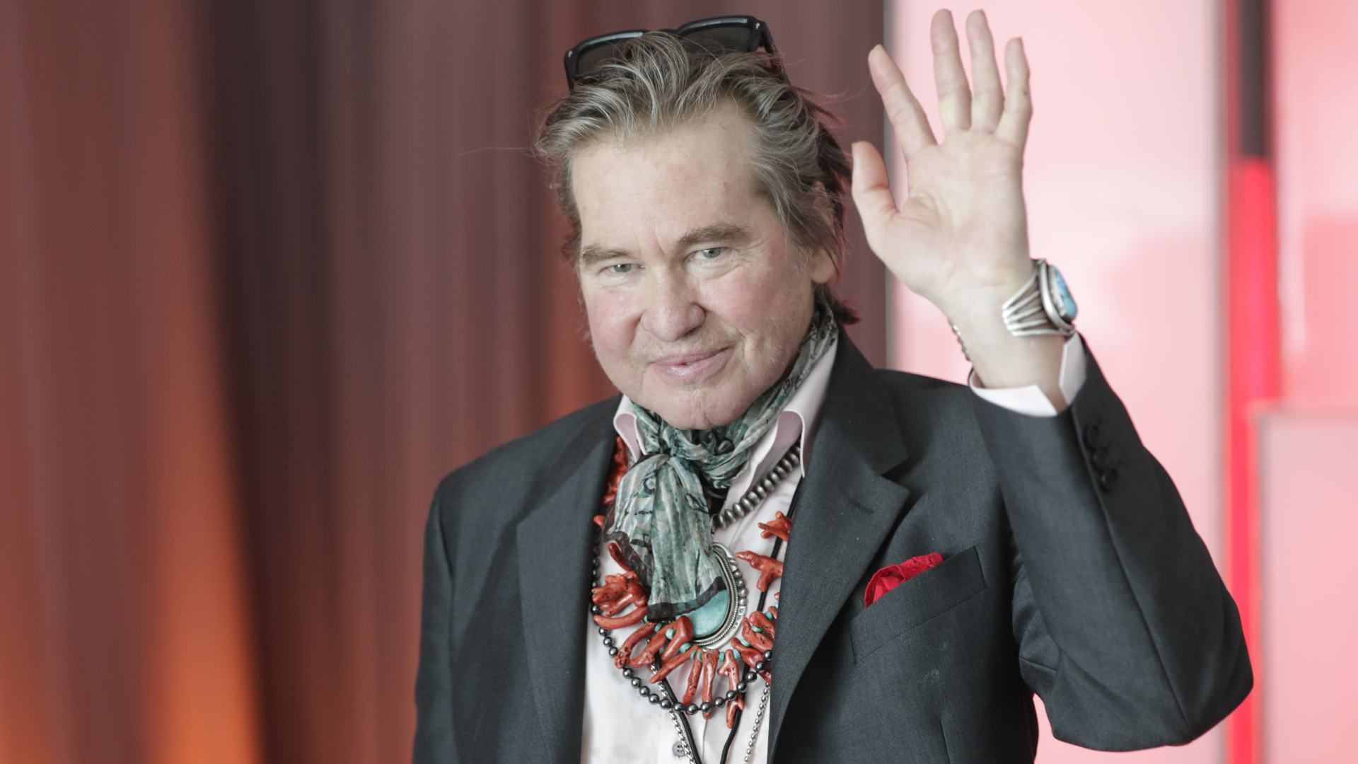 Val Kilmer waves, wearing a suit, scarf, and sunglasses on his head, with a necklace and watch.