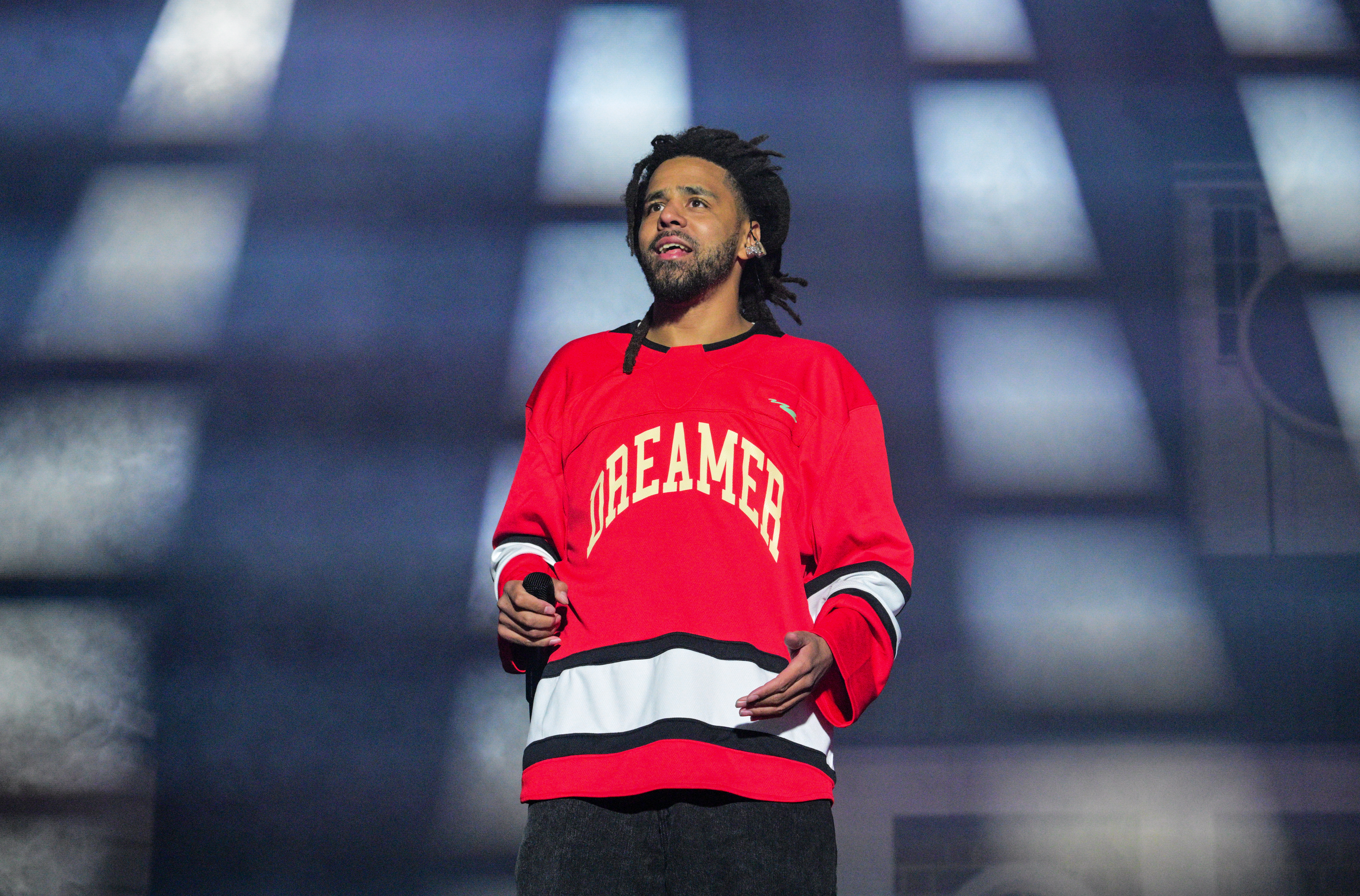 J. Cole on stage wearing a red "Dreamer" jersey, holding a microphone, with a backdrop of lights.