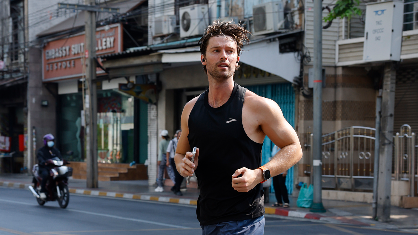 A man jogging on a city street, wearing a black tank top and holding a phone. Buildings and a motorbike are in the background.