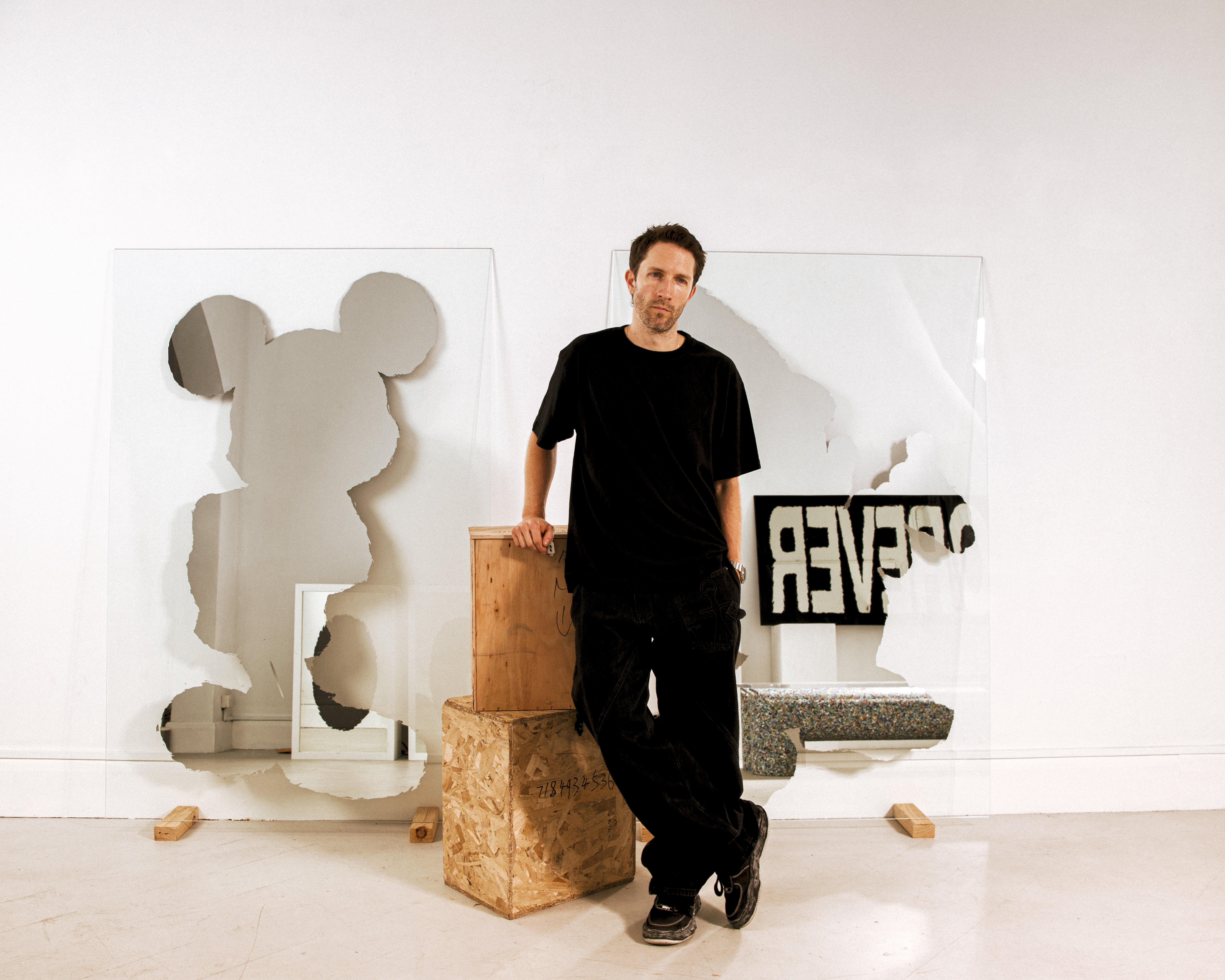 Man in black clothing stands in an art studio with Mickey Mouse and "FOREVER" cutouts on glass panels.