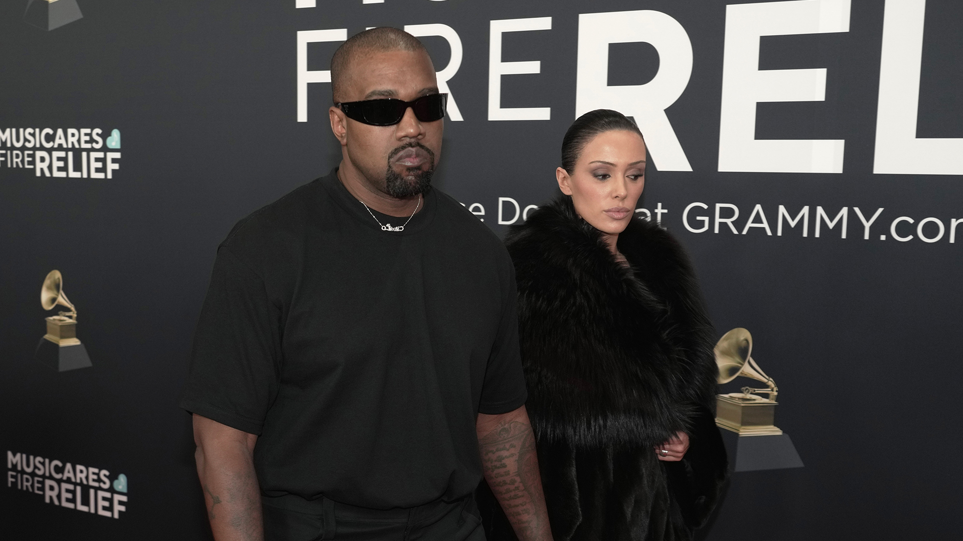 Kanye West in sunglasses and black attire, accompanied by a Bianca Censori in a fur coat, at a MusiCares event backdrop.