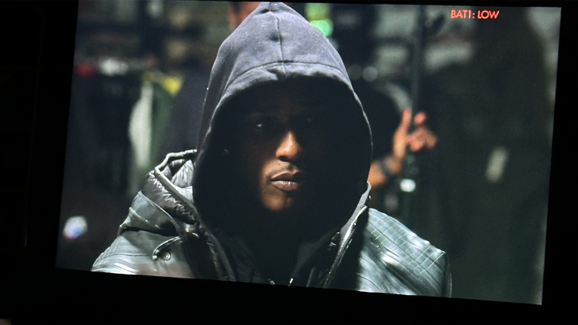 ASAP Rocky wearing a dark hooded jacket is sitting in a dimly lit environment, looking serious and focused.