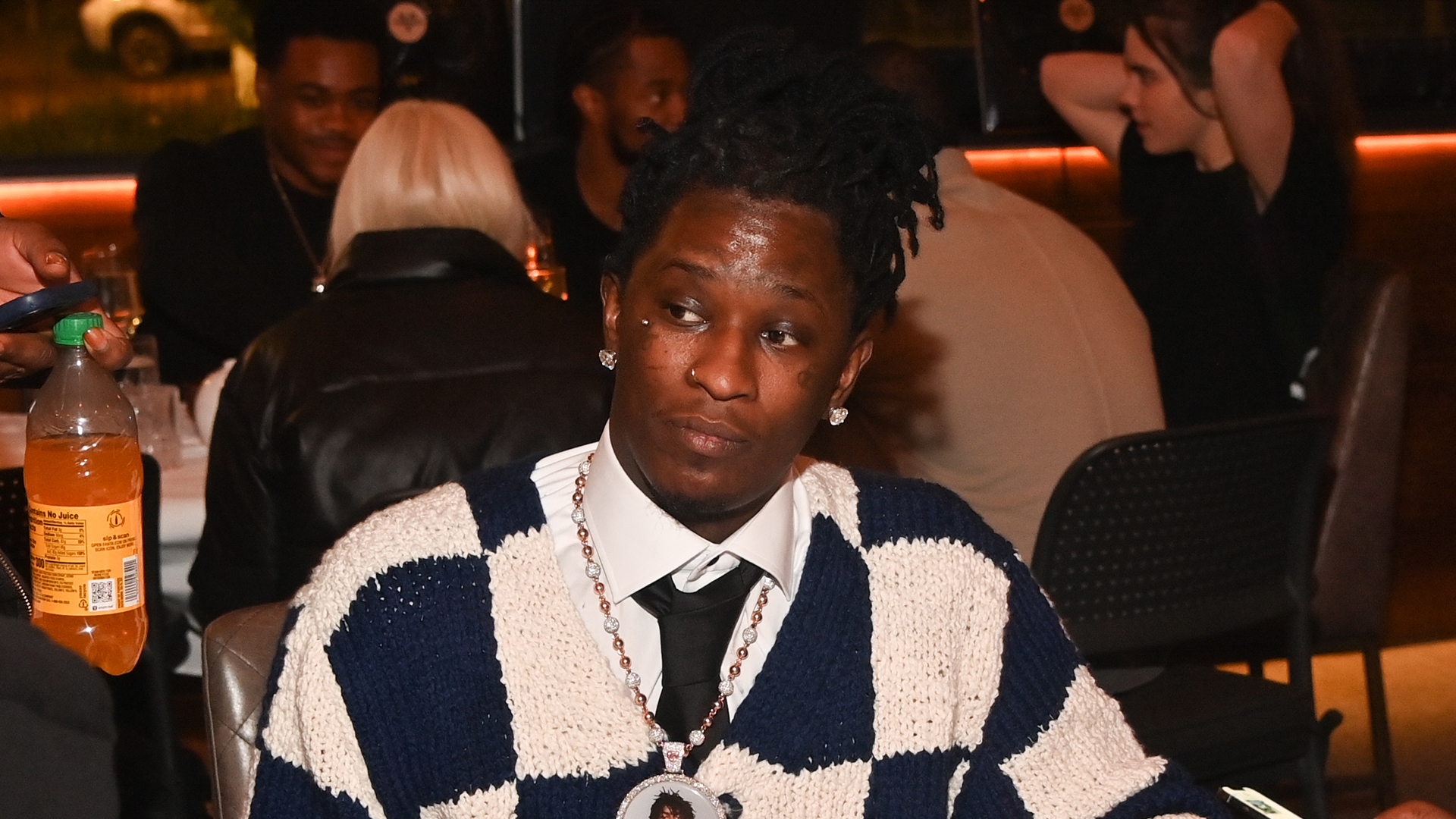 Young Thug in a checkered sweater and tie sits at a table in a restaurant, surrounded by people.