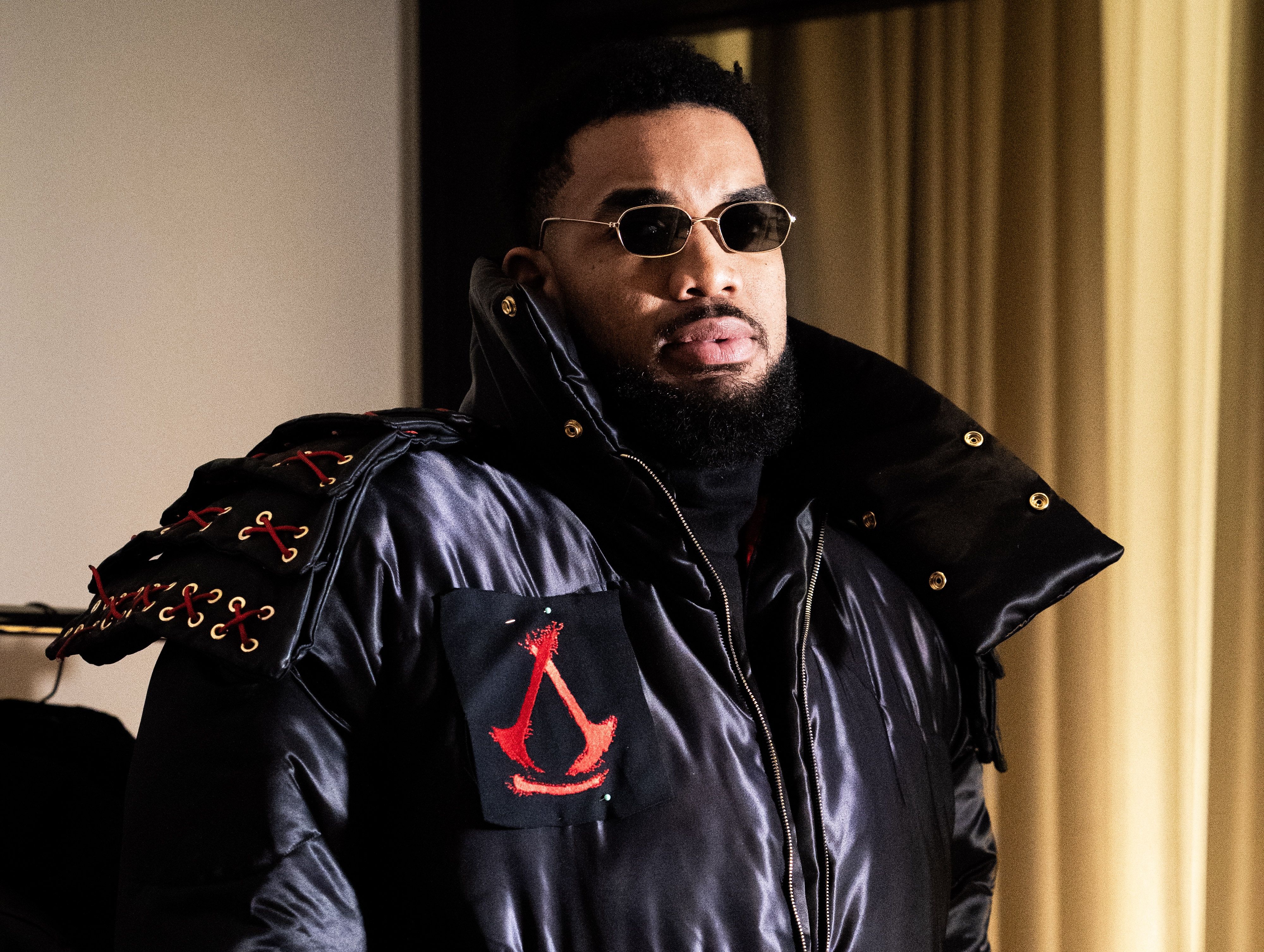 A man wearing sunglasses and a black jacket with red accents stands indoors.
