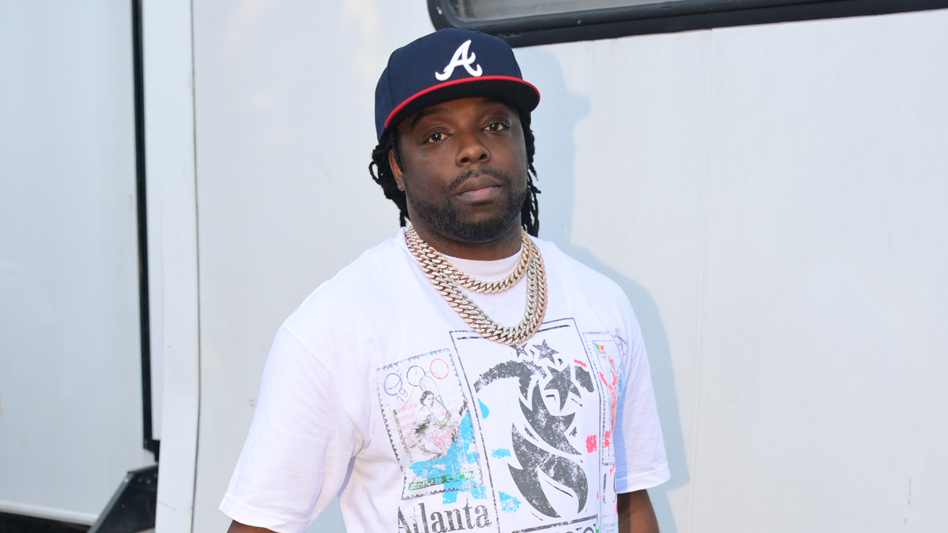 Young Scooter wearing an Atlanta Braves cap, white graphic t-shirt, and gold chains stands in front of a light-colored background.
