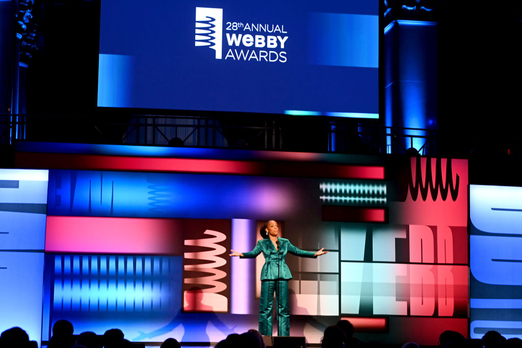 A person in a teal suit stands on stage at the 28th Annual Webby Awards, with colorful abstract designs in the background.