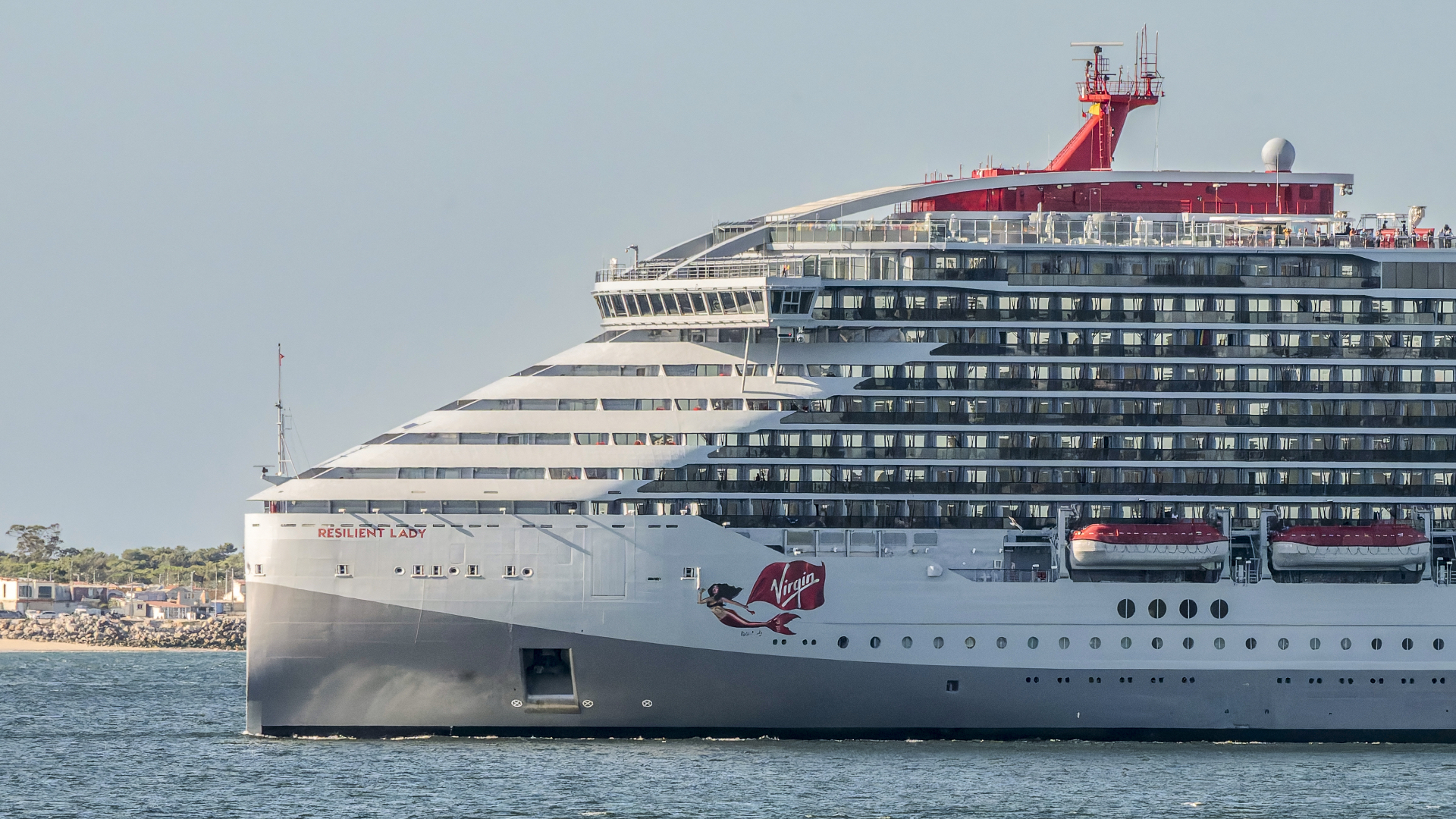 A large cruise ship, the "Resilient Lady" by Virgin Voyages, sails on the water with multiple decks and lifeboats visible.