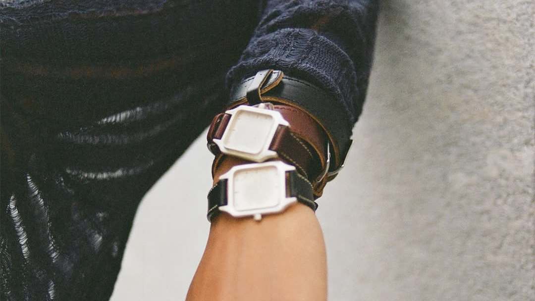 A person wearing two watches with square faces and leather straps on their wrist, against a textured background. Our Legacy