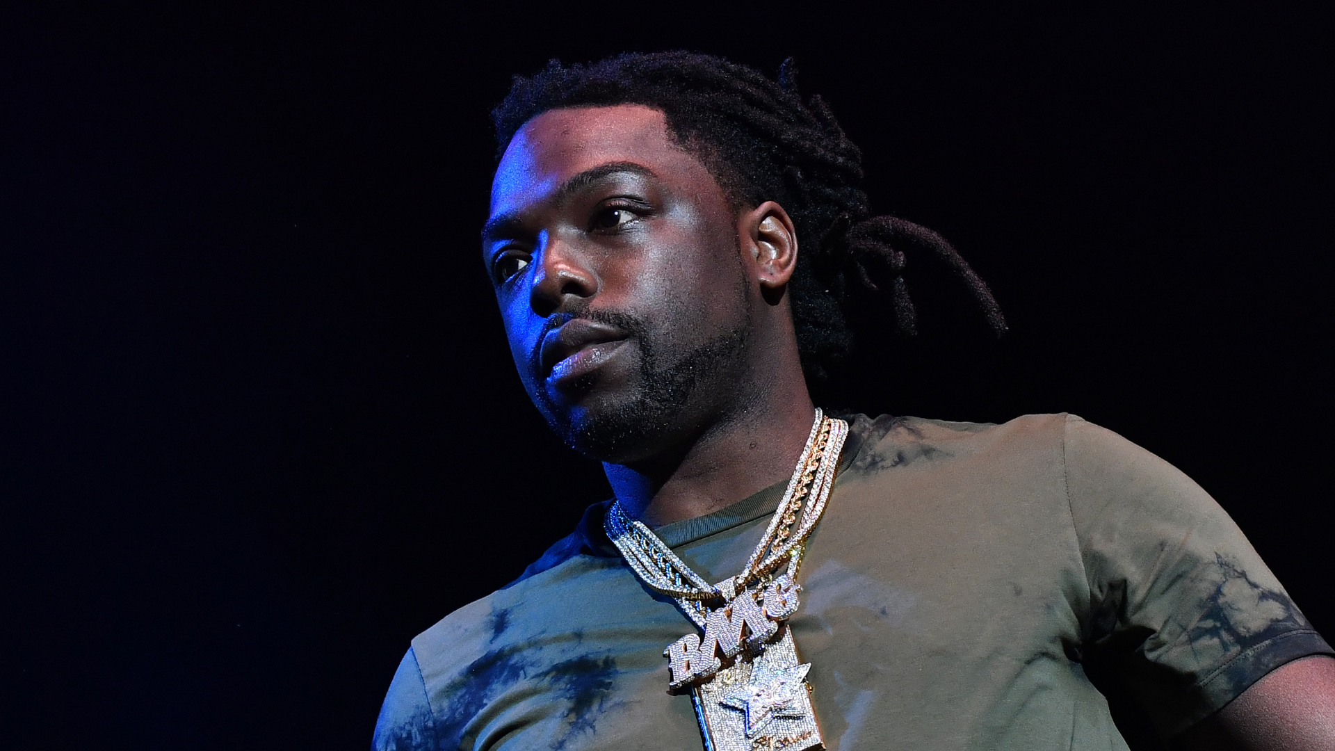 A man with dreadlocks wearing a green shirt and layered necklaces with large pendants, set against a dark background.