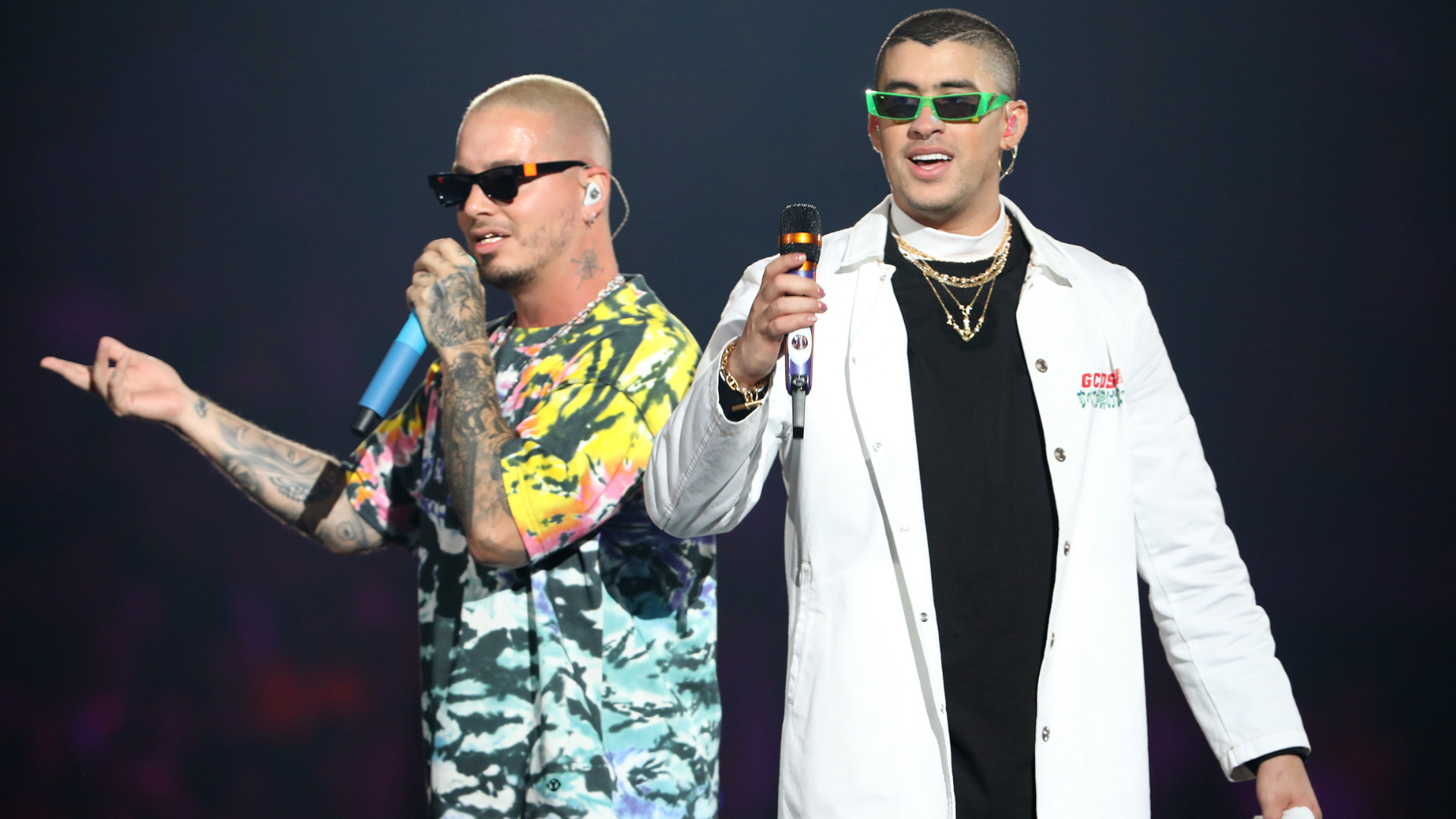 J Balvin and Bad Bunny performing on stage; J Balvin in a colorful shirt and Bad Bunny in a white coat with green sunglasses.