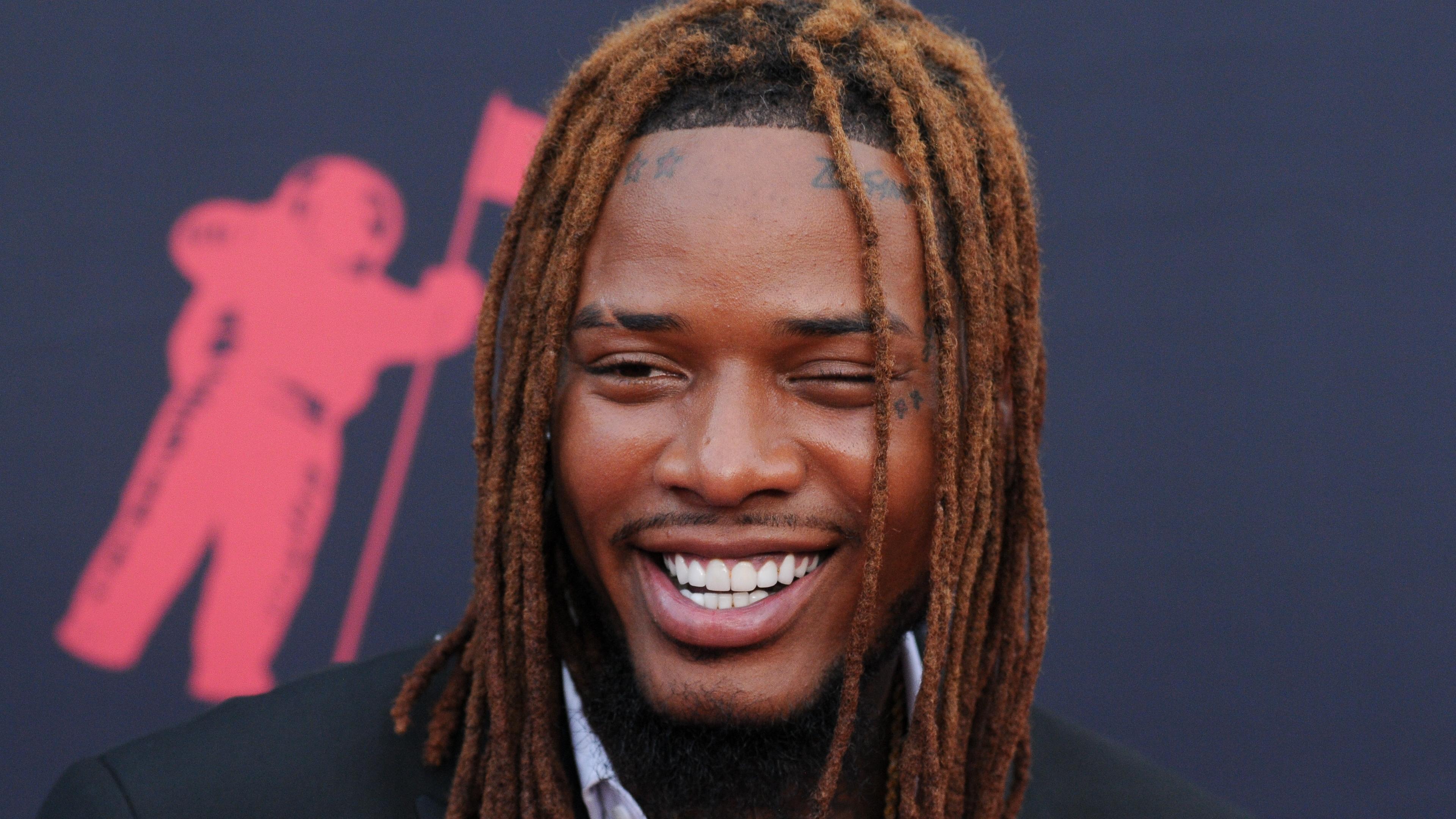 Fetty Wap smiling on a red carpet, wearing a black suit and white shirt, with dreadlocks and tattoos visible.