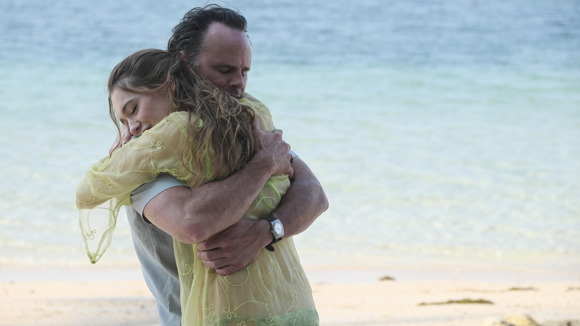 Chelsea and Rick from 'The White Lotus' embrace on a beach, with the ocean in the background. Chelsea wears a light green dress, and Rick wears a watch.