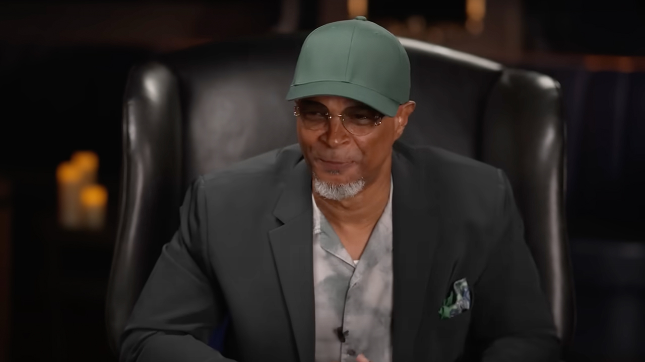 Damon Wayans Sr. sits on a black chair, wearing a gray blazer and patterned shirt, with a subtle smile.