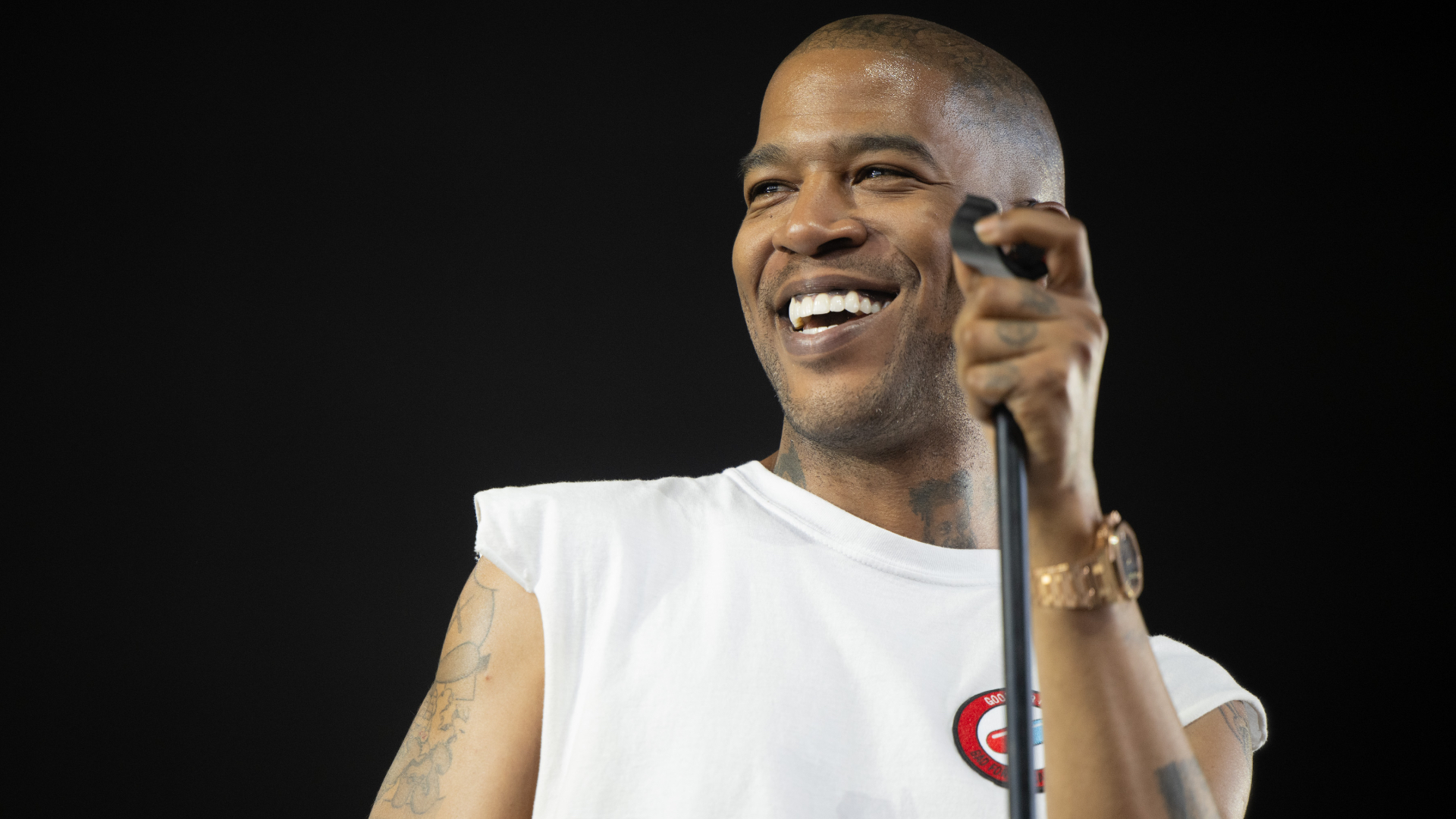 Kid Cudi smiling while holding a microphone, wearing a white sleeveless shirt and a watch, performing on stage.