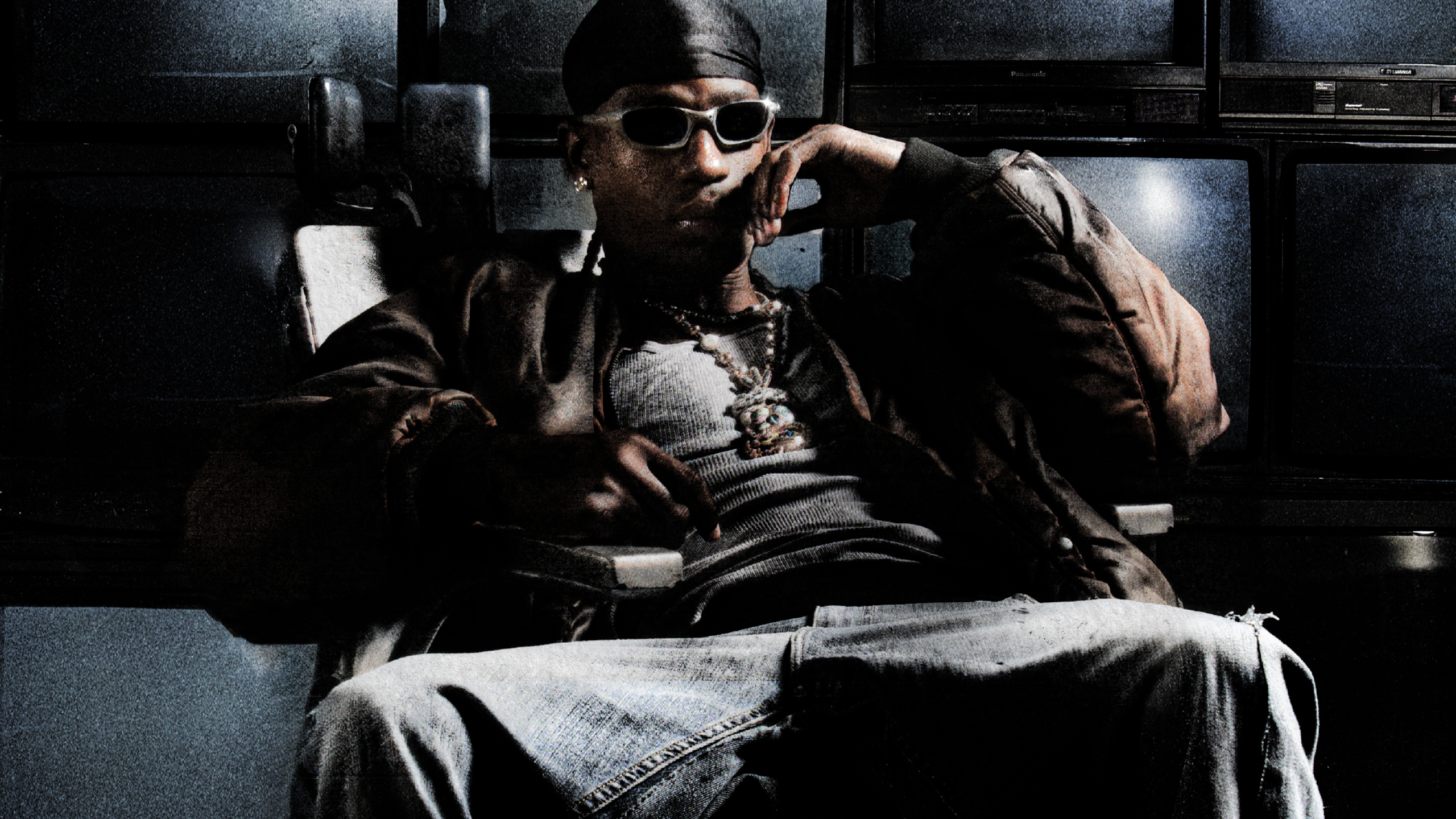 A person wearing sunglasses and a bandana sits in a chair, dressed in a dark jacket and jeans, with multiple TV screens in the background.