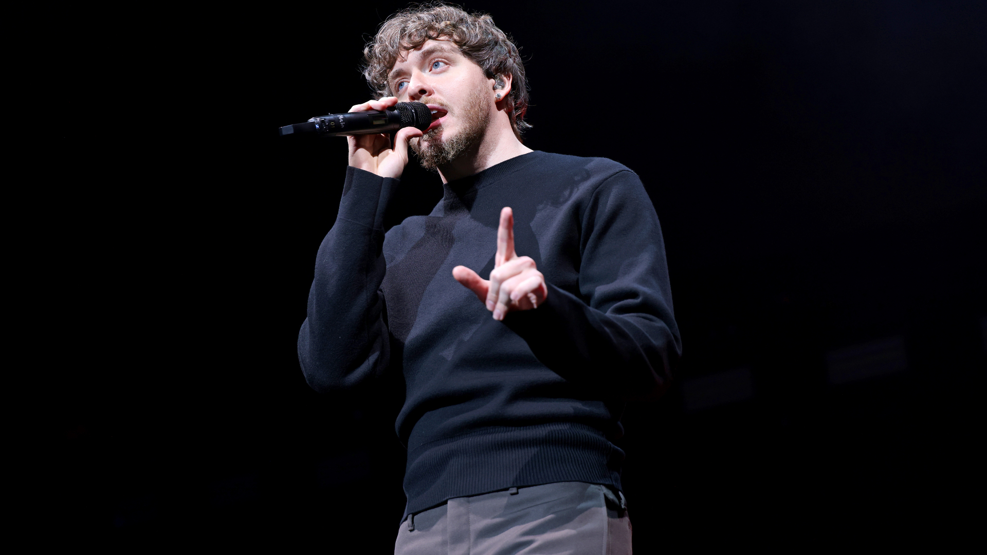 Jack Harlow with curly hair and a beard, wearing a black sweater, is singing into a microphone on stage.